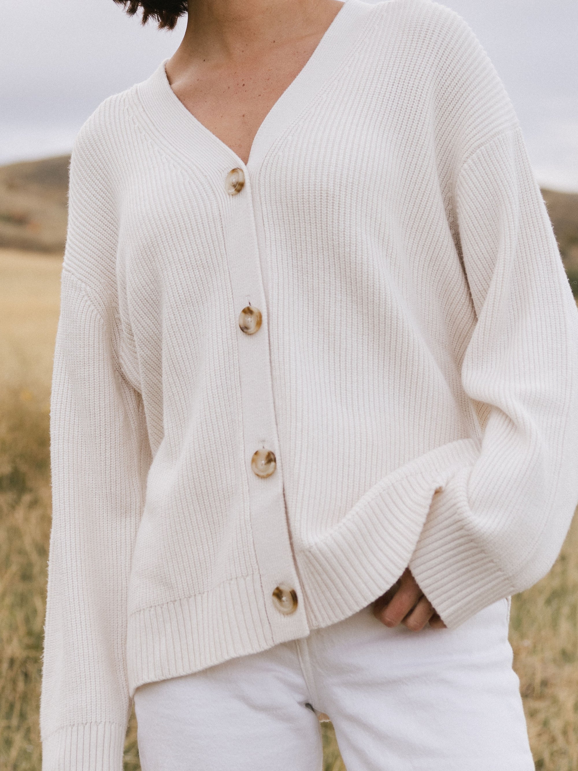 A woman wearing Cozy Earth Classic Cardigan in white stands in a field. She is pictured from the neck down 