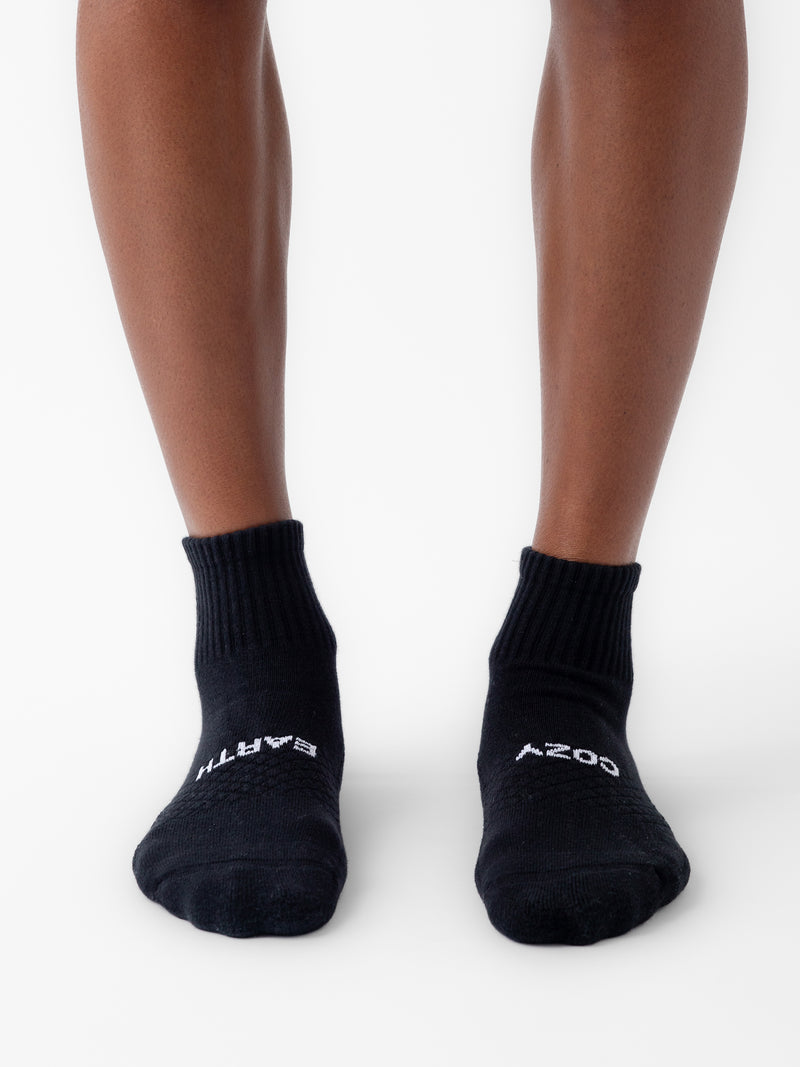 A person stands with feet slightly apart, wearing black ankle socks from the Essential Quarter Sock 2-Pack by Cozy Earth. The socks feature white text—"EARTH" on the left sock and "COSY" on the right sock. The background is plain white.