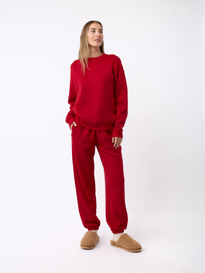 A person stands against a plain background, wearing a matching red sweatshirt and sweatpants set. They have light brown hair and are gazing to the side. On their feet, they are wearing Cozy Earth's Puffy Sheep Slippers in tan, which look fuzzy and comfortable. They have one hand in their pocket.
