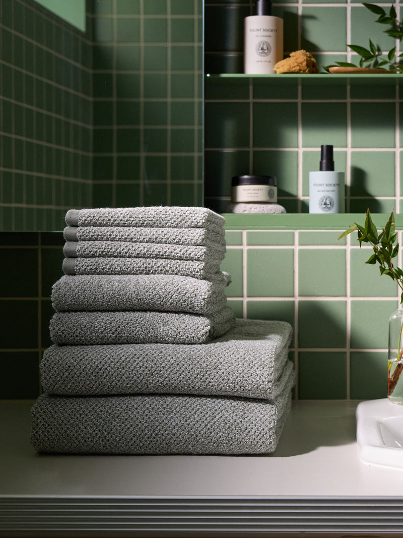 A stack of Nantucket Towels in Heathered Sage is sitting on the countertop of a bathroom vanity. A small plant and shelves of Fount Society skincare products are shown in the background.
