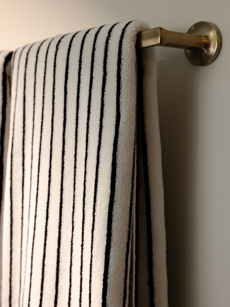 A Luxe Bath Towel from Cozy Earth, featuring elegant black vertical stripes on a pristine white fabric, hangs neatly on a brass towel rod mounted on a white wall. Soft lighting casts gentle shadows, accentuating the towel's plush texture.