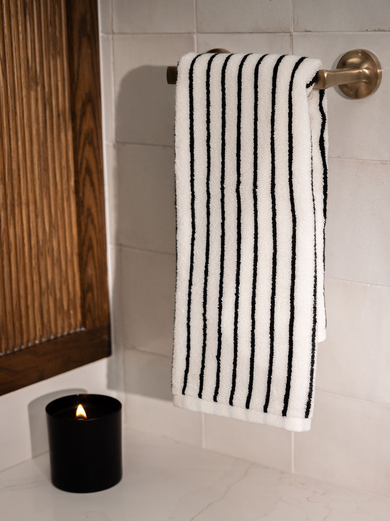 A neatly folded Luxe Hand Towel by Cozy Earth, featuring a white fabric with black vertical stripes, hangs on a silver towel rack mounted on a tiled wall. Below, a black candle container with a lit flame sits on a white countertop next to a wooden decor element.