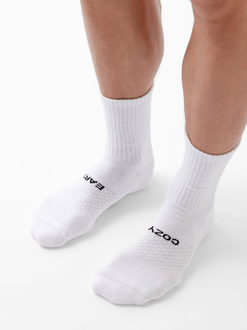 A person stands on a white surface wearing Cozy Earth’s Essential Calf Sock 8-Pack in white. The left sock reads "EARTH" and the right reads "COZY" in black letters near the toes.