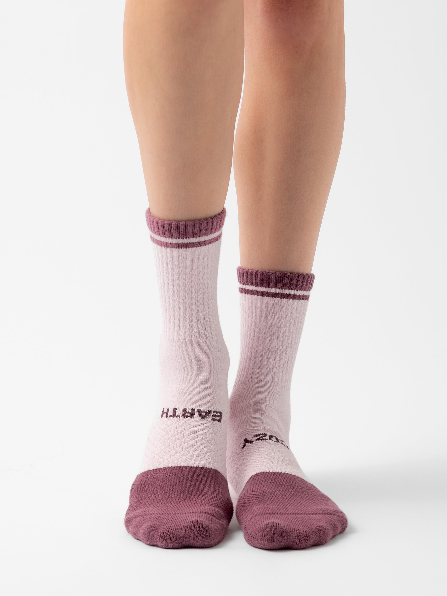 A person wearing mismatched pink socks with maroon accents from the Essential Calf Sock 4-Pack by Cozy Earth. The left sock displays the word "EARTH" on the side, while the right one features "COZY." These socks have ribbed tops and toes, and the photo is set against a plain white background. |Color:Arctic/Lilac