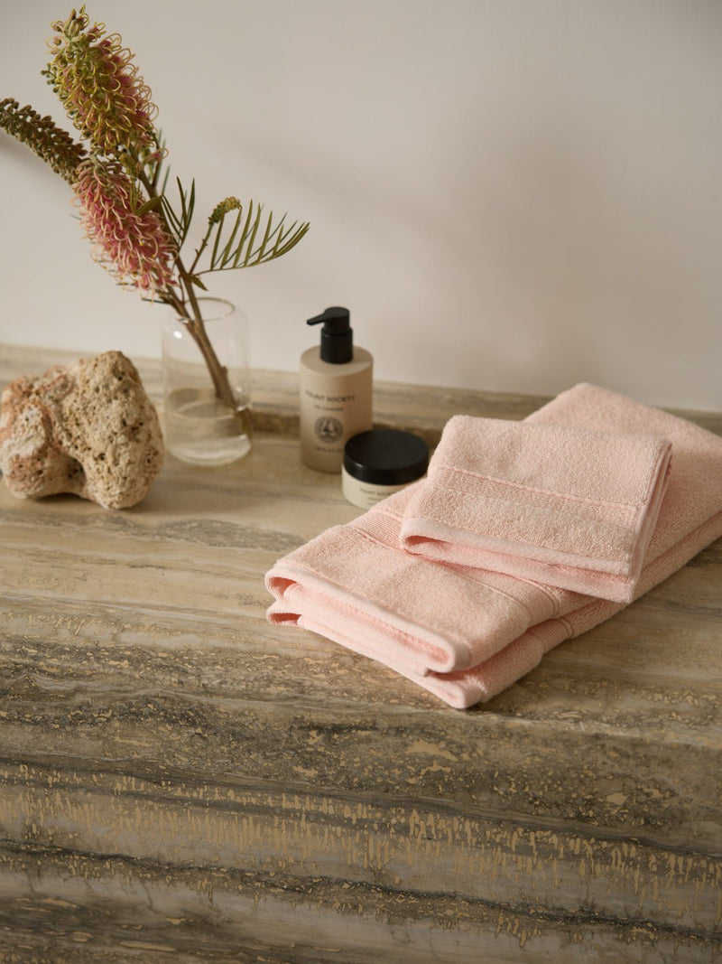 Cozy Earth's Luxe Hand Towels in pink are folded on a marble surface beside a small plant in a glass vase, a large stone, a pump bottle, and a closed jar. The neutral tones and minimalist decor create a soothing atmosphere.