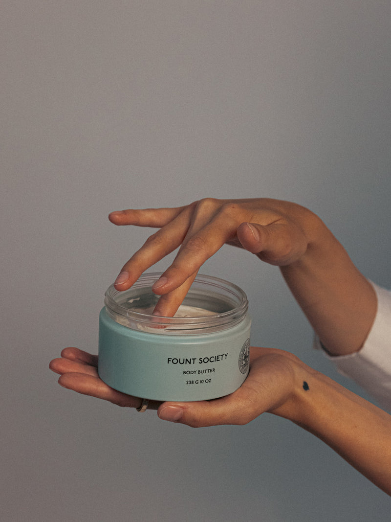 Two hands appear in the image; one holds an open Dewy Duo jar from Cozy Earth, while fingers from the other hand dip into the cream. The jar is light blue with black text set against a neutral backdrop.