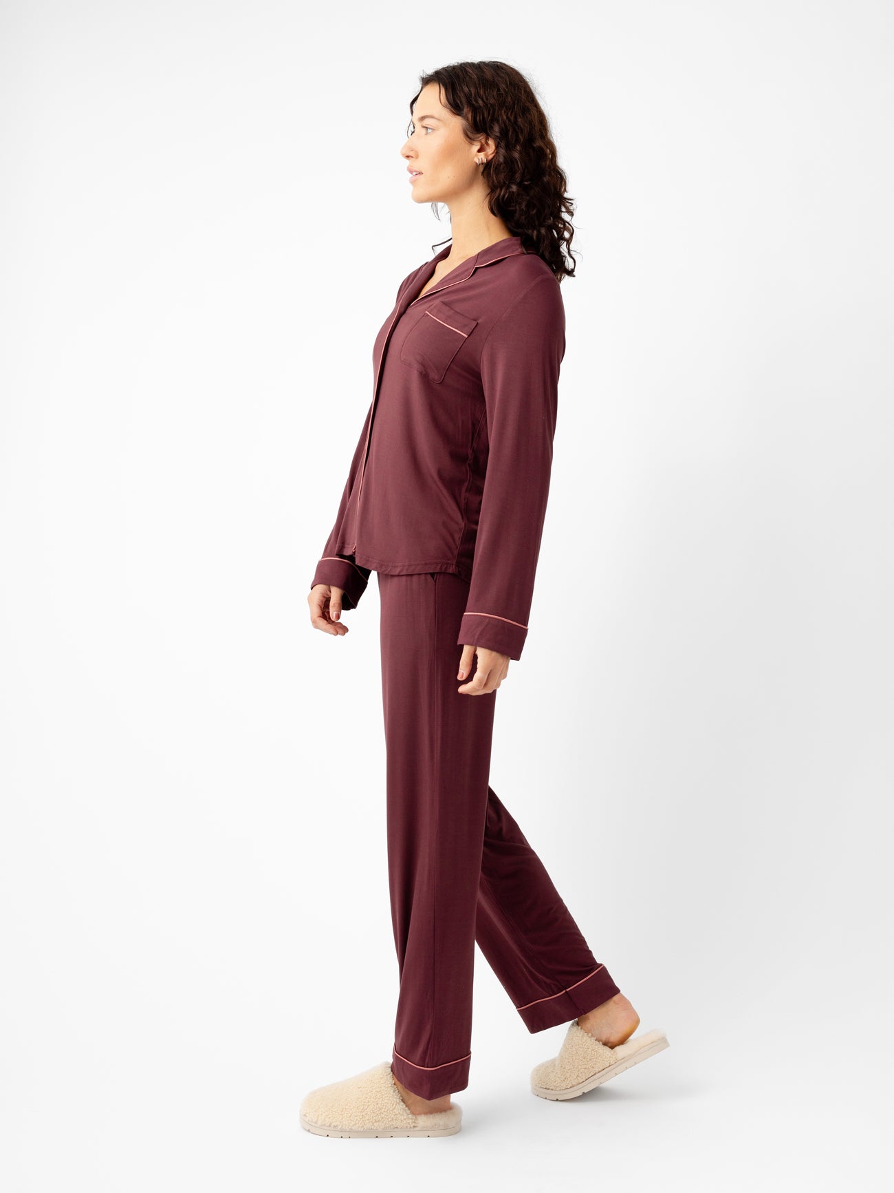 A person with wavy hair stands in profile against a plain white background, wearing the Last Chance Women's Bamboo Stretch-Knit Long Sleeve Pajama Set by Cozy Earth in maroon and beige slippers. 
