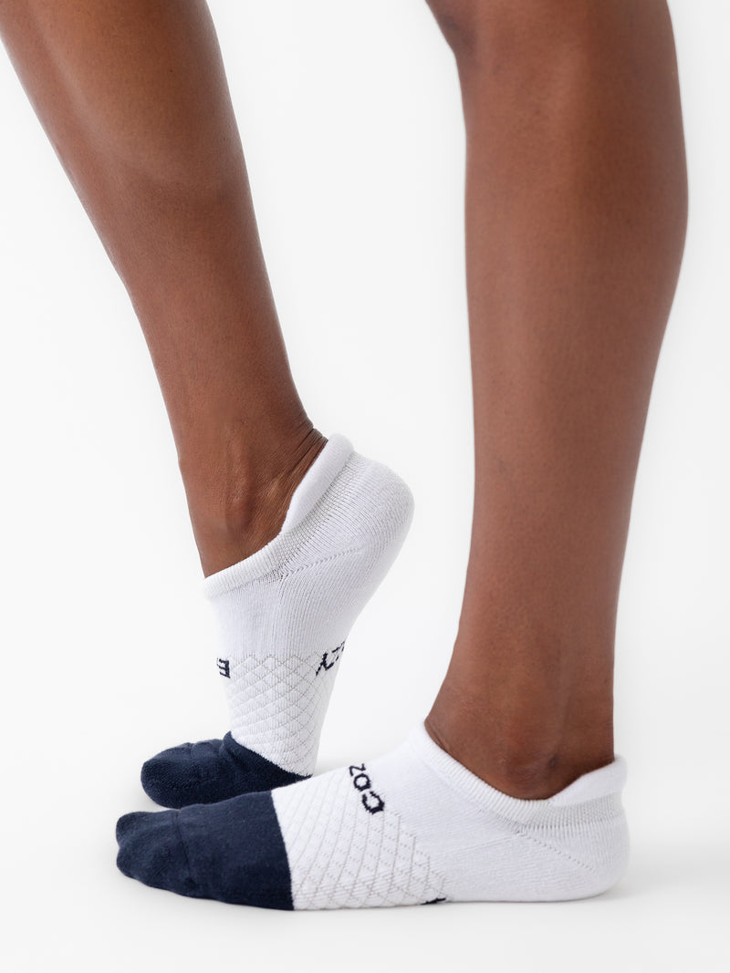Close-up of two feet wearing Cozy Earth Essential Ankle Socks from a 2-pack, featuring white fabric with black toes and heels. The person stands on tiptoe against a plain white background, highlighting the socks' snug fit and cushioned bottoms.
