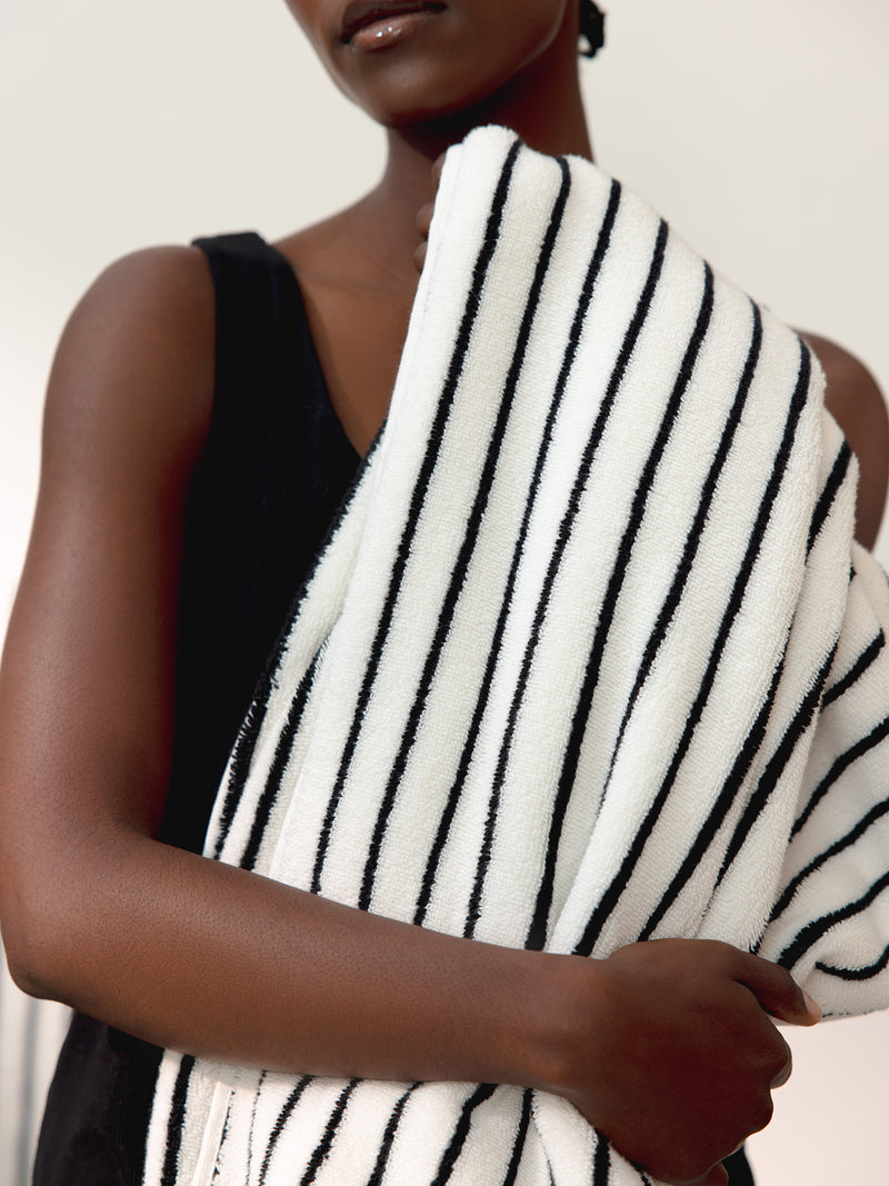 A person wearing a black sleeveless top holds a Luxe Bath Towel by Cozy Earth, featuring black stripes on white fabric, draped over one shoulder. The background is blurred and neutral. The person's face is partially shown from the nose down.