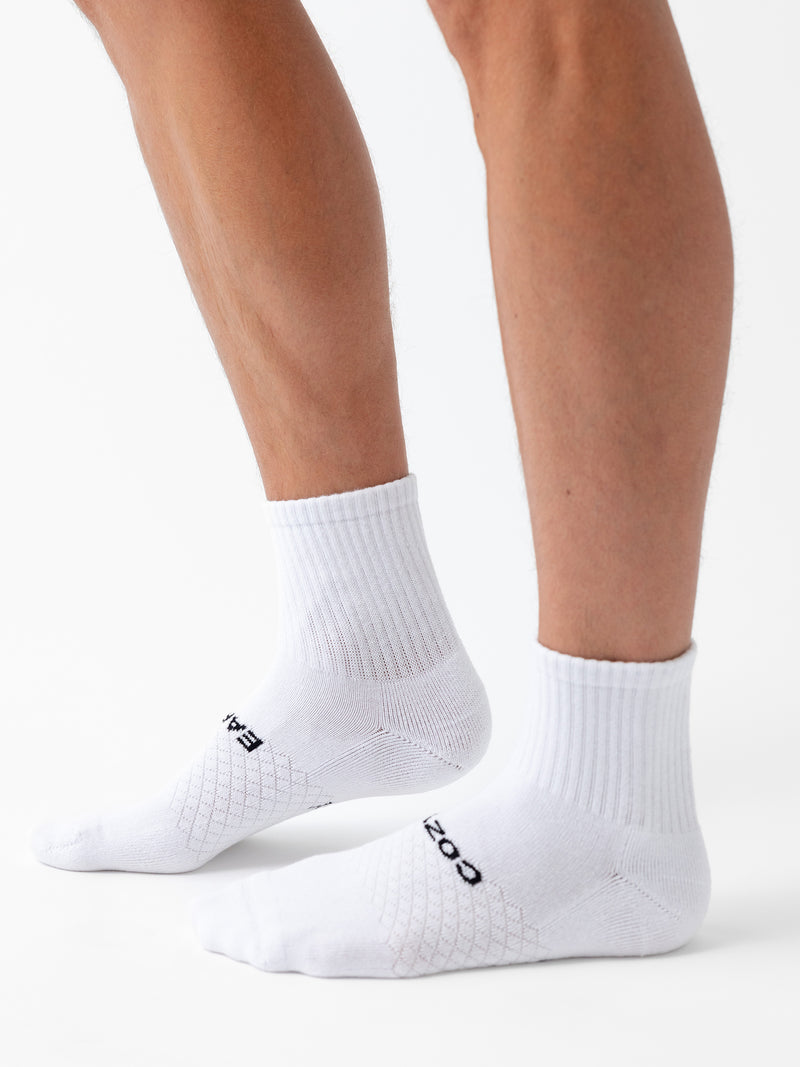 A person wearing Cozy Earth Essential Quarter Sock 6-Pack, featuring white socks with black text on top, stands against a white background. Only their lower legs and feet are visible.
