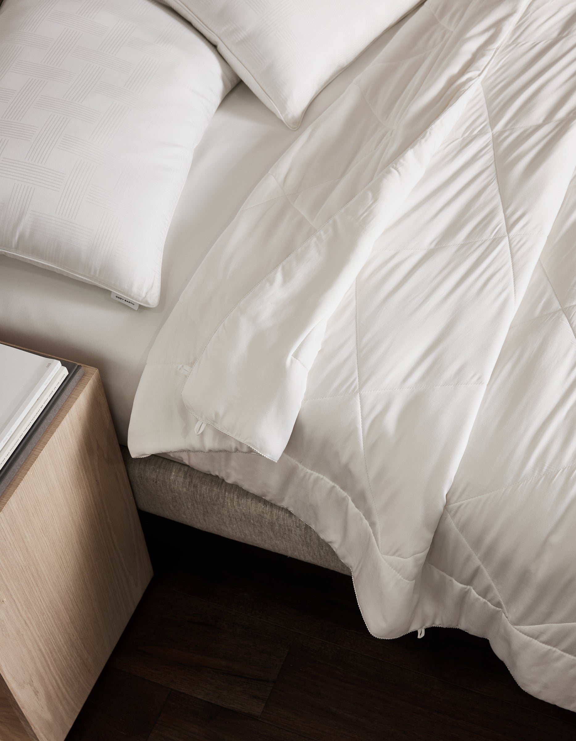 A bed with Cozy Earth's Bamboo Insert Bundle—white pillows and a quilted comforter—beside a light wood nightstand on a dark wood floor.
