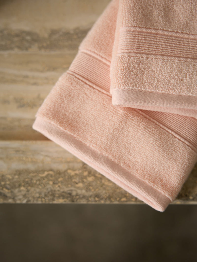Two folded Luxe Hand Towels from Cozy Earth, in a peach color, rest on a textured wooden surface. Their soft texture and simple design are showcased against an earthy-toned background, complementing their gentle hue.