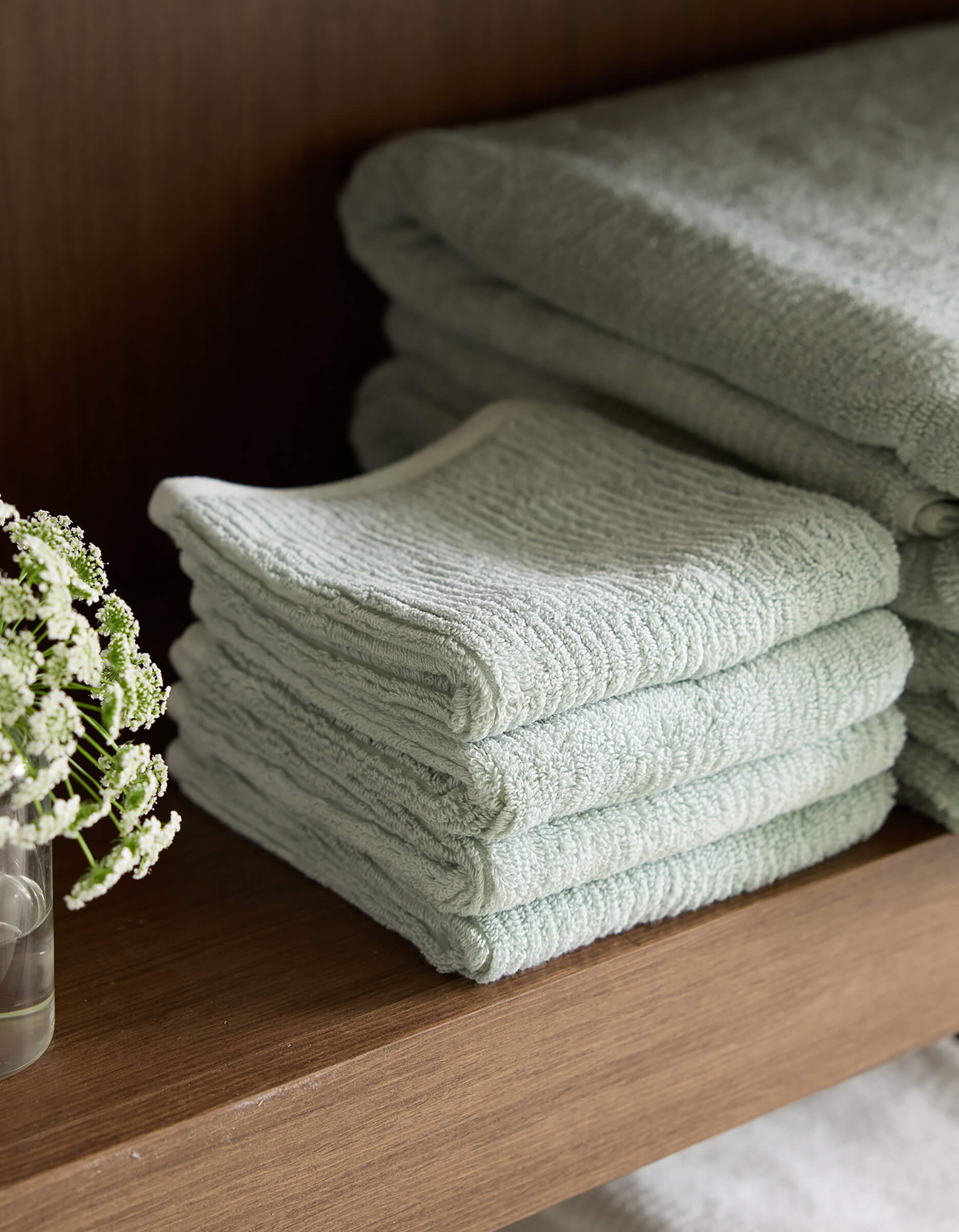 A neatly folded stack of Cozy Earth's Ribbed Terry Washcloths in pale green is placed on a wooden shelf. Beside the washcloths, a small clear glass with a few delicate white flowers in water adds a touch of elegance to the display. 