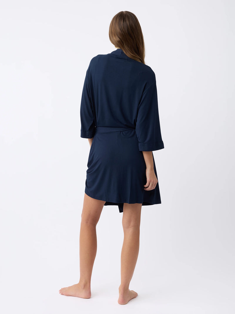 A person with long hair stands barefoot on a plain white background, facing away while wearing Cozy Earth's Women's Stretch-Knit Bamboo Kimono Robe in dark blue with three-quarter sleeves.