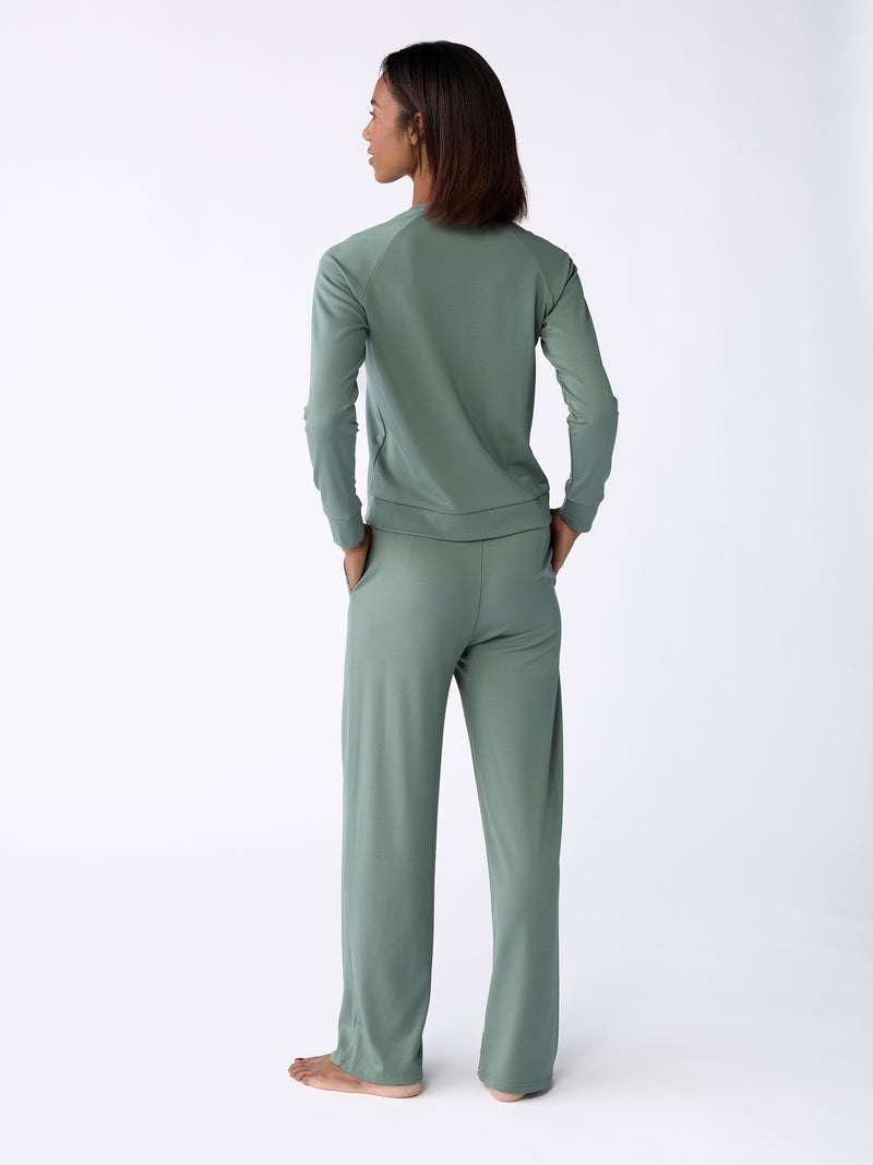 A person stands barefoot, facing away while wearing the Women's Ultra-Soft Bamboo Wide Leg Pull On Pant TALL in sage green from Cozy Earth. The background is a plain, light color.