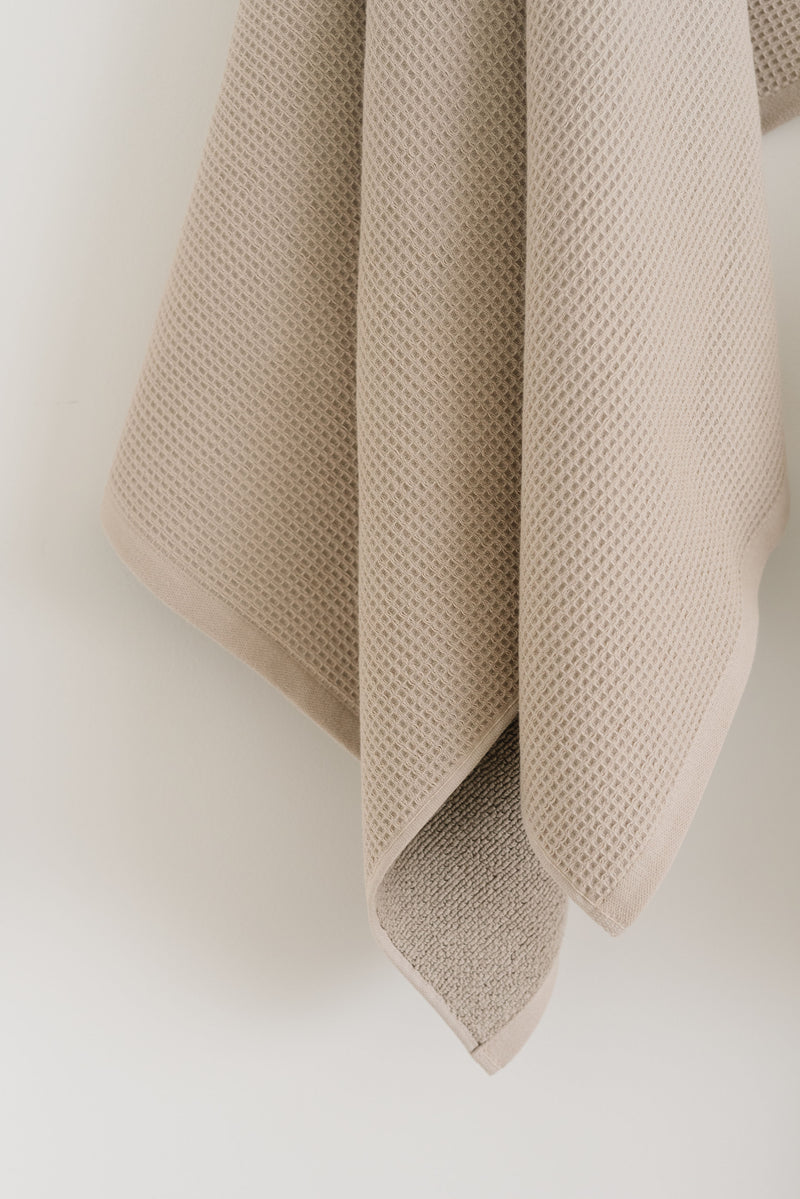 Waffle Bath Towel in the color Sand. Photo of Sand Waffle Bath Towel taken hanging from a towel hook in a white bathroom. The upper half of the towel and towel hook is not seen.