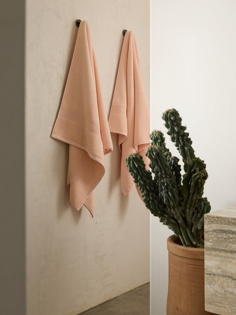 Cozy Earth's Luxe Bath Sheets in soft pink hang on a light-colored wall. A tall, twisted potted cactus adds greenery to the minimalist backdrop, with a stone countertop partially visible on the right.