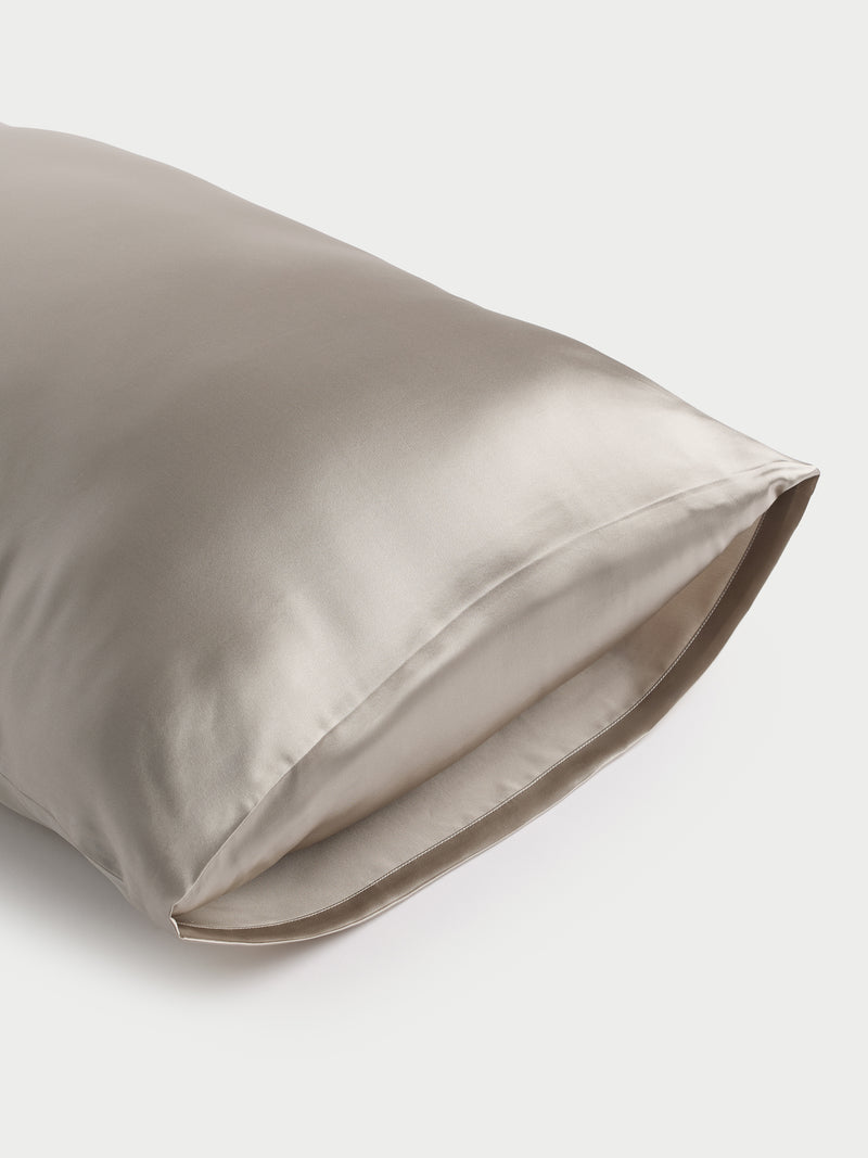 A smooth, shimmering silver Cozy Earth Silk Pillowcase is displayed against a plain white background. The fabric looks incredibly soft and luxurious, with a subtle sheen indicating its silky texture. One end of the pillowcase is slightly open.