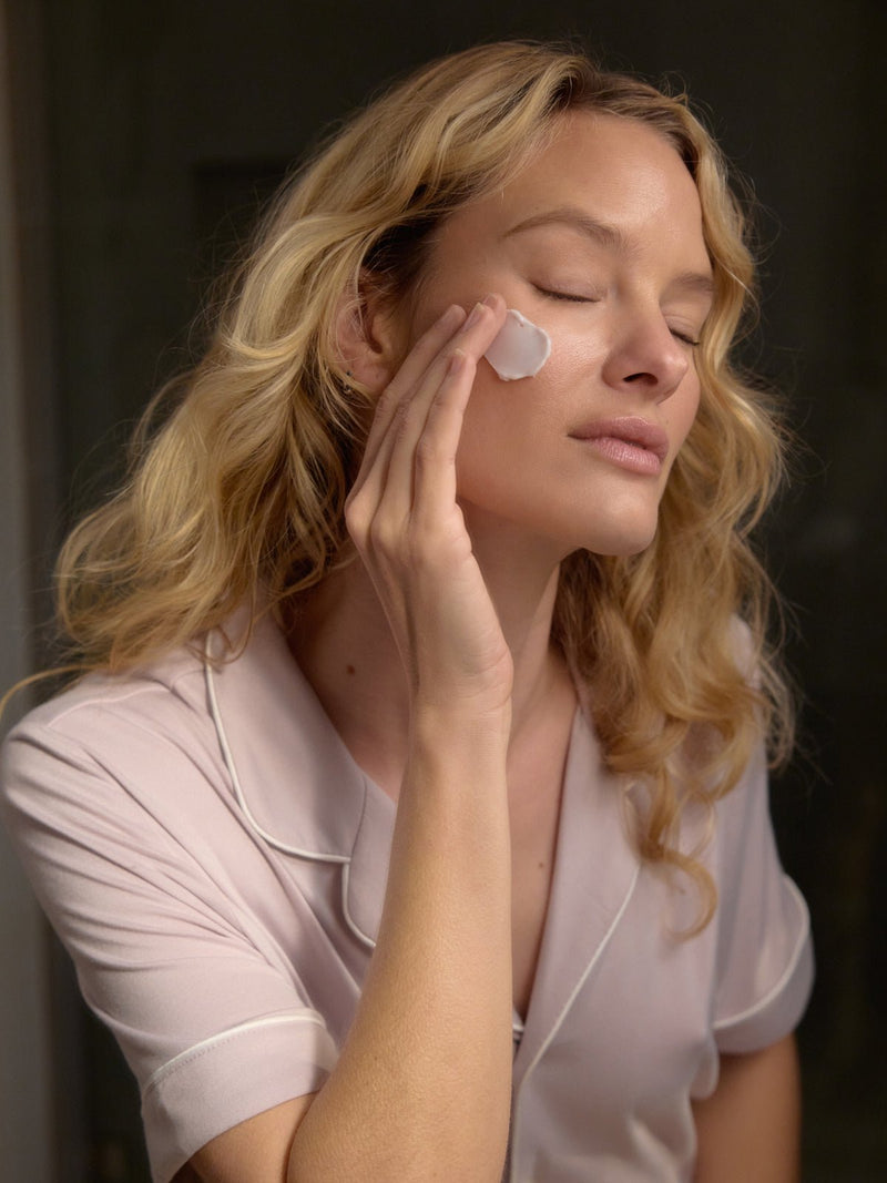 A woman with long blonde hair gently applies Cozy Earth's Night Cream to her cheek with her fingers. Her eyes are softly closed, and she appears serene and relaxed in a light-colored top against a softly blurred background.