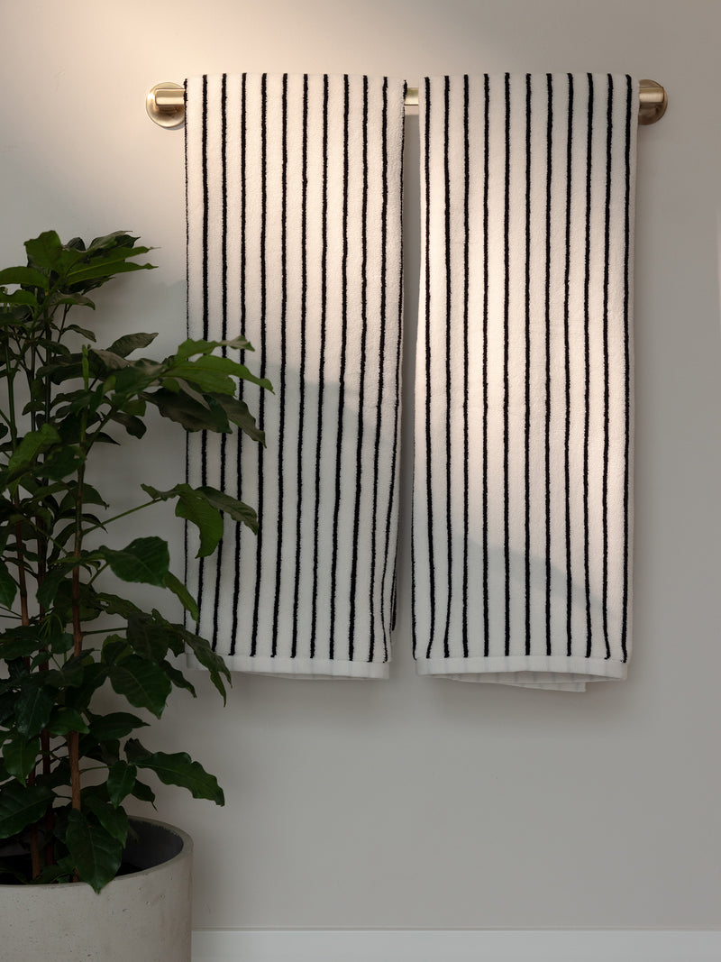 Two Luxe Bath Towels by Cozy Earth, featuring black vertical stripes on a white background, hang neatly on a golden towel rack attached to a light-colored wall. A potted green plant sits on the floor to the left of the towels, adding a touch of nature to the scene.