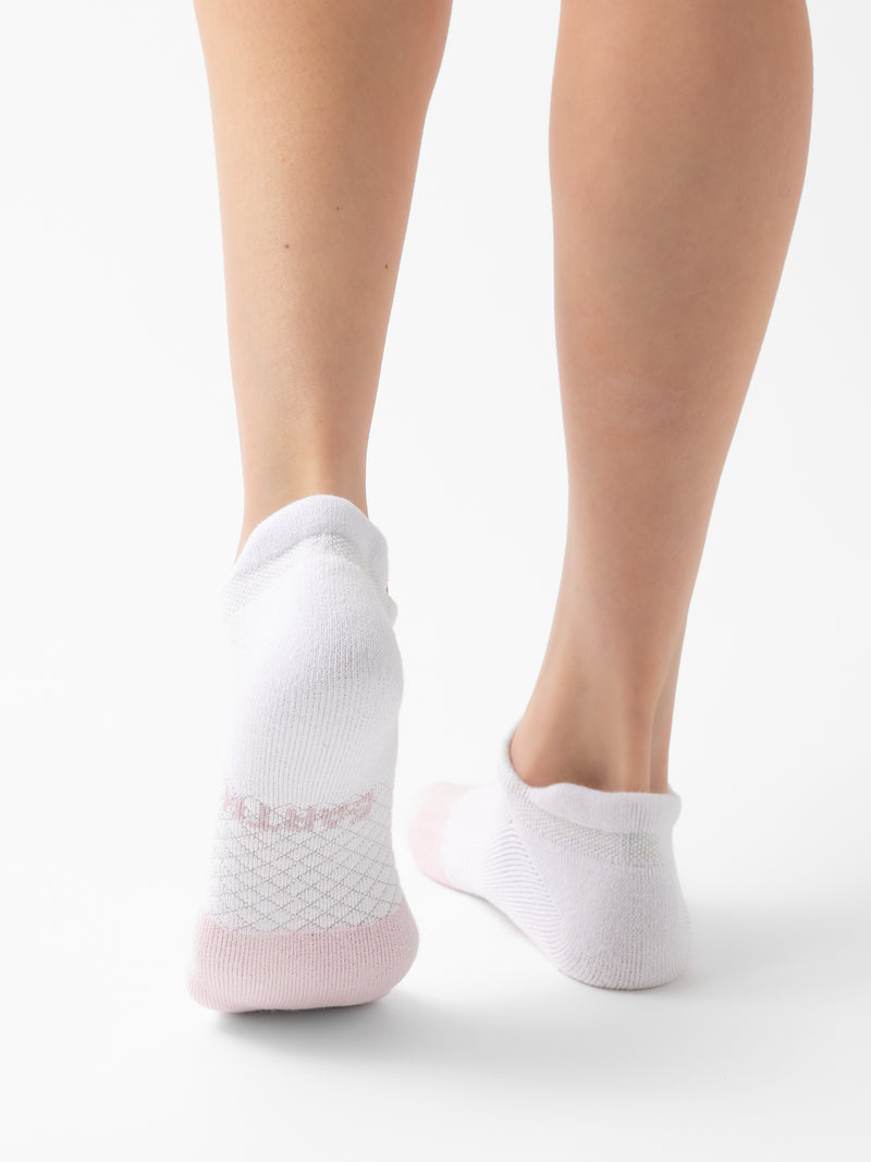 A person standing on a white surface, wearing a pair of Cozy Earth's Essential Ankle Sock 2-Pack in white. The socks feature a textured pattern around the heel and sole areas. Only the lower legs and feet are visible from the back.