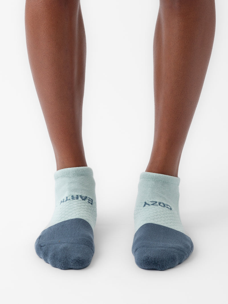 A pair of feet wearing light blue ankle socks from the Cozy Earth Essential Ankle Sock 2-Pack, featuring dark blue toes and heels. One sock displays the word "EARTH" while the other reads "COZY.