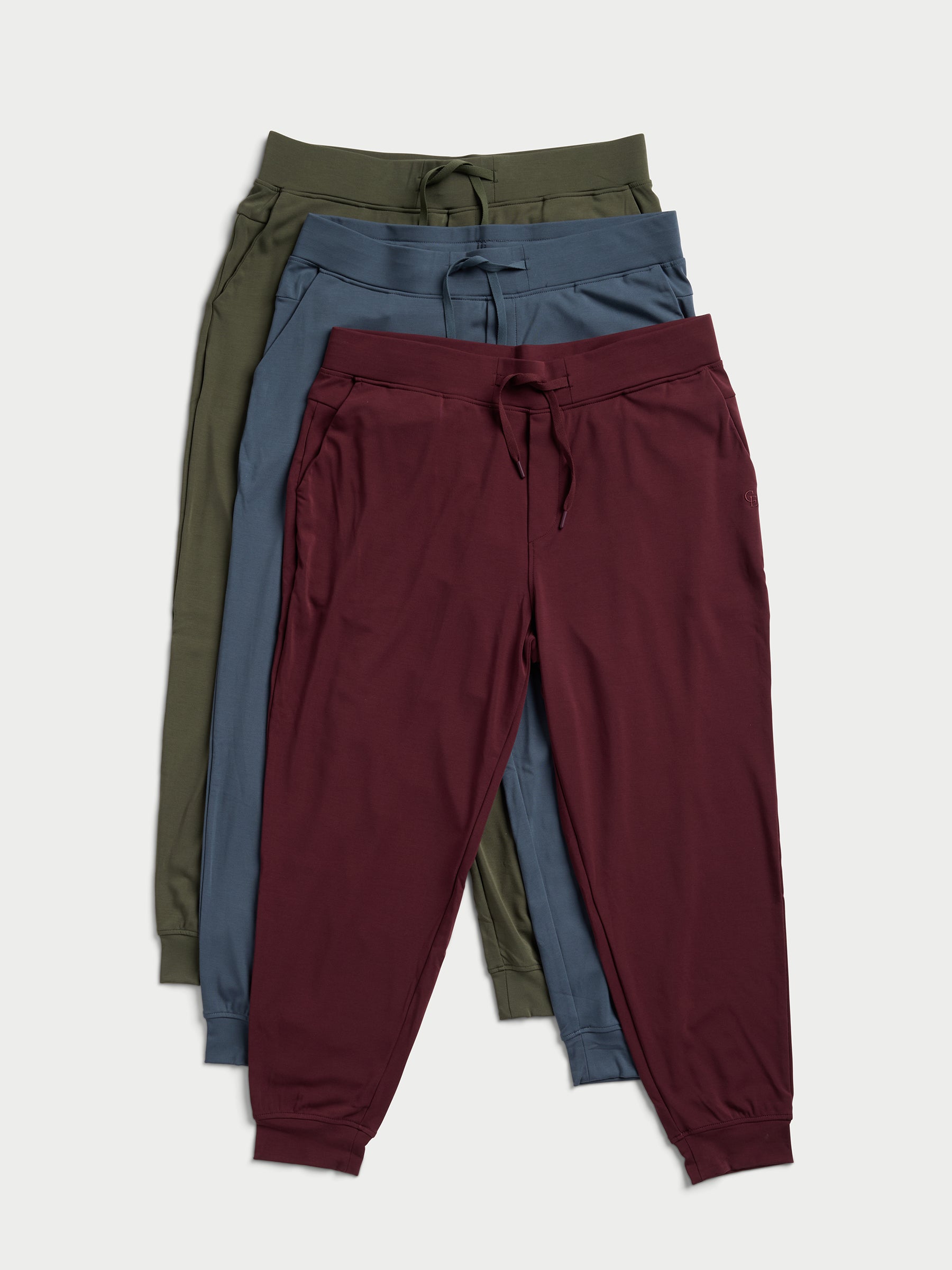 There are three Men's Ultra-Soft Bamboo Pullover Crew pants by Cozy Earth stacked in different colors: green on top, blue in the middle, and maroon at the bottom, each featuring drawstrings at the waist and cuffs at the ankles. 