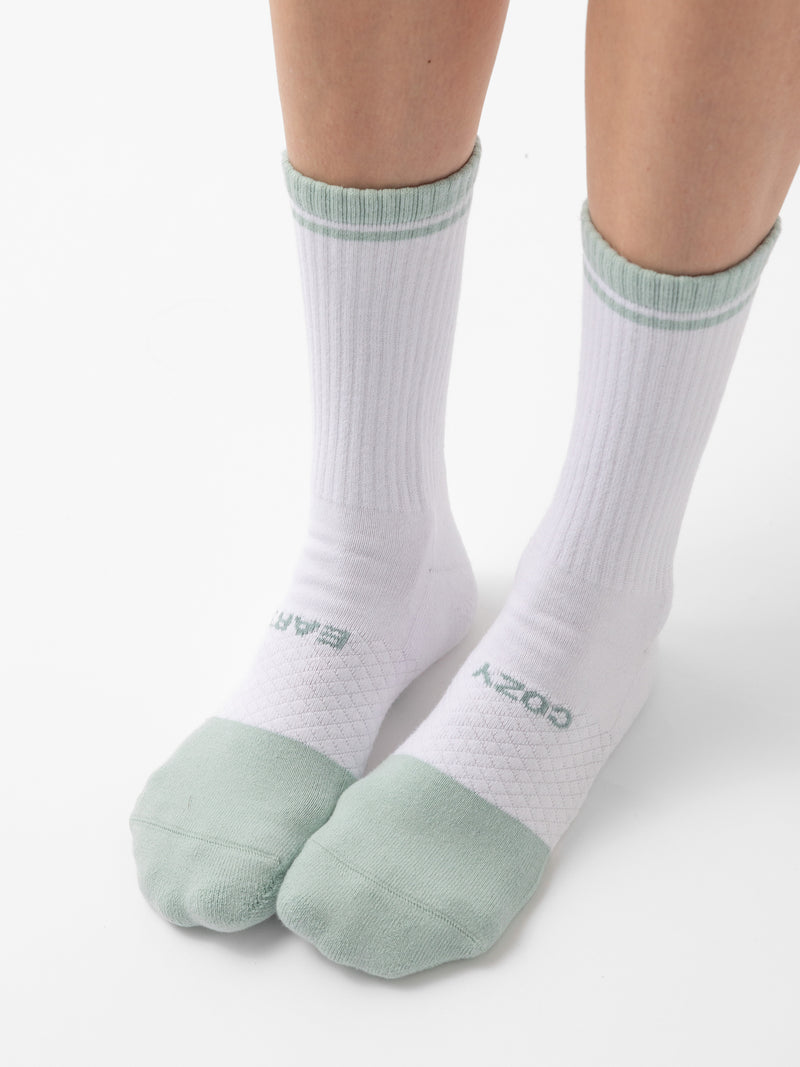 Two feet are wearing white socks with light green toes, heels, and top bands. The socks feature a ribbed section and have the "Cozy Earth" brand name written near the bottom. The background is plain white. These are part of the Essential Calf Sock 4-Pack collection by Cozy Earth.
