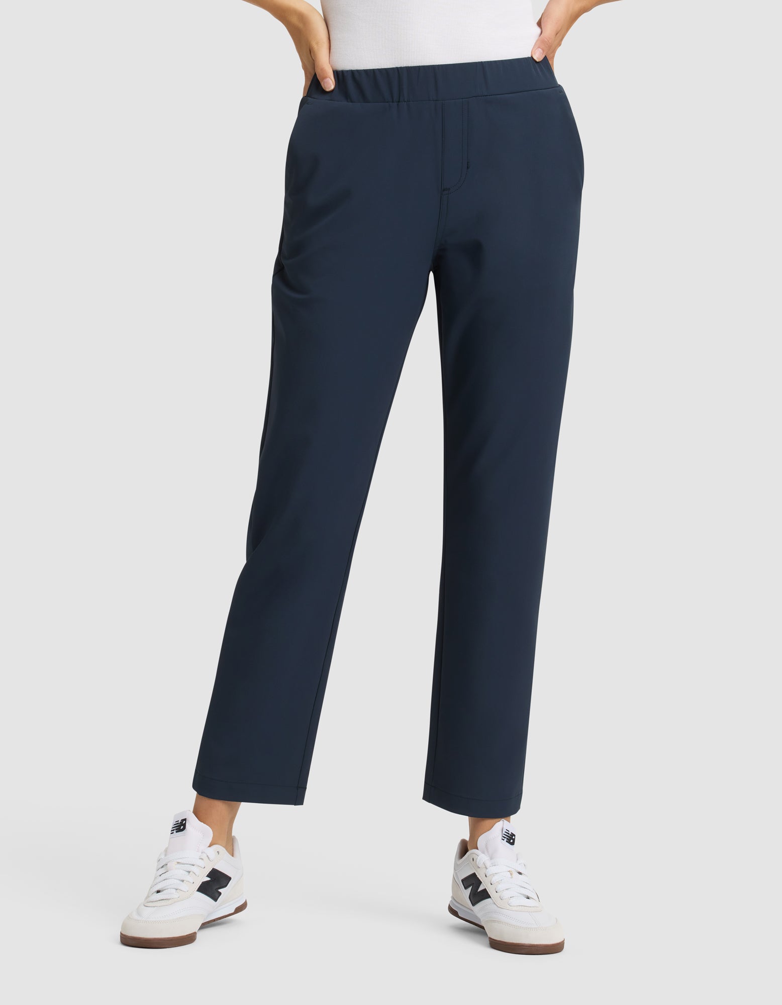 A person stands against a plain light background wearing Cozy Earth's Women's Always Cropped Pant in navy blue with an elastic waistband, paired with a white top and white New Balance sneakers. Their hands are in their pockets. 