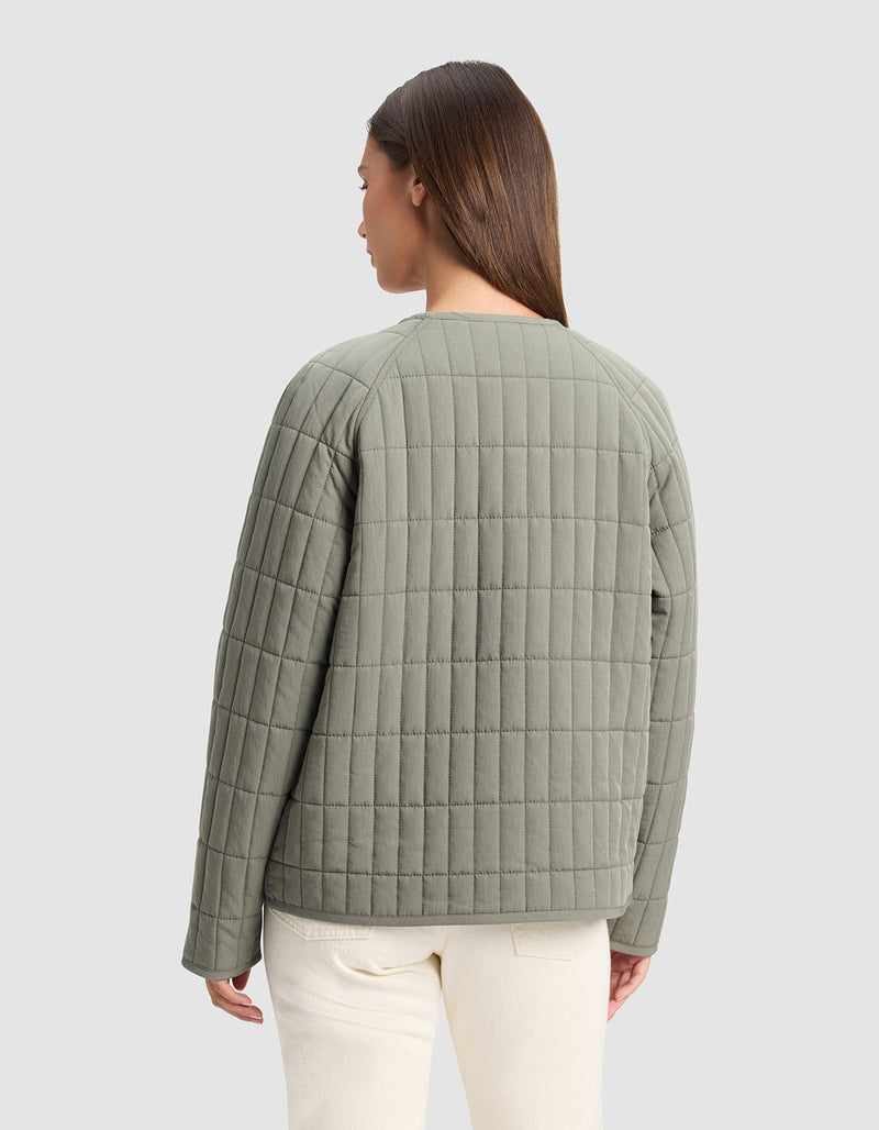 A woman with straight brown hair, facing away from the camera, wears the Cozy Earth Women's Andie Quilted Jacket in light olive green and off-white pants against a plain light gray background.