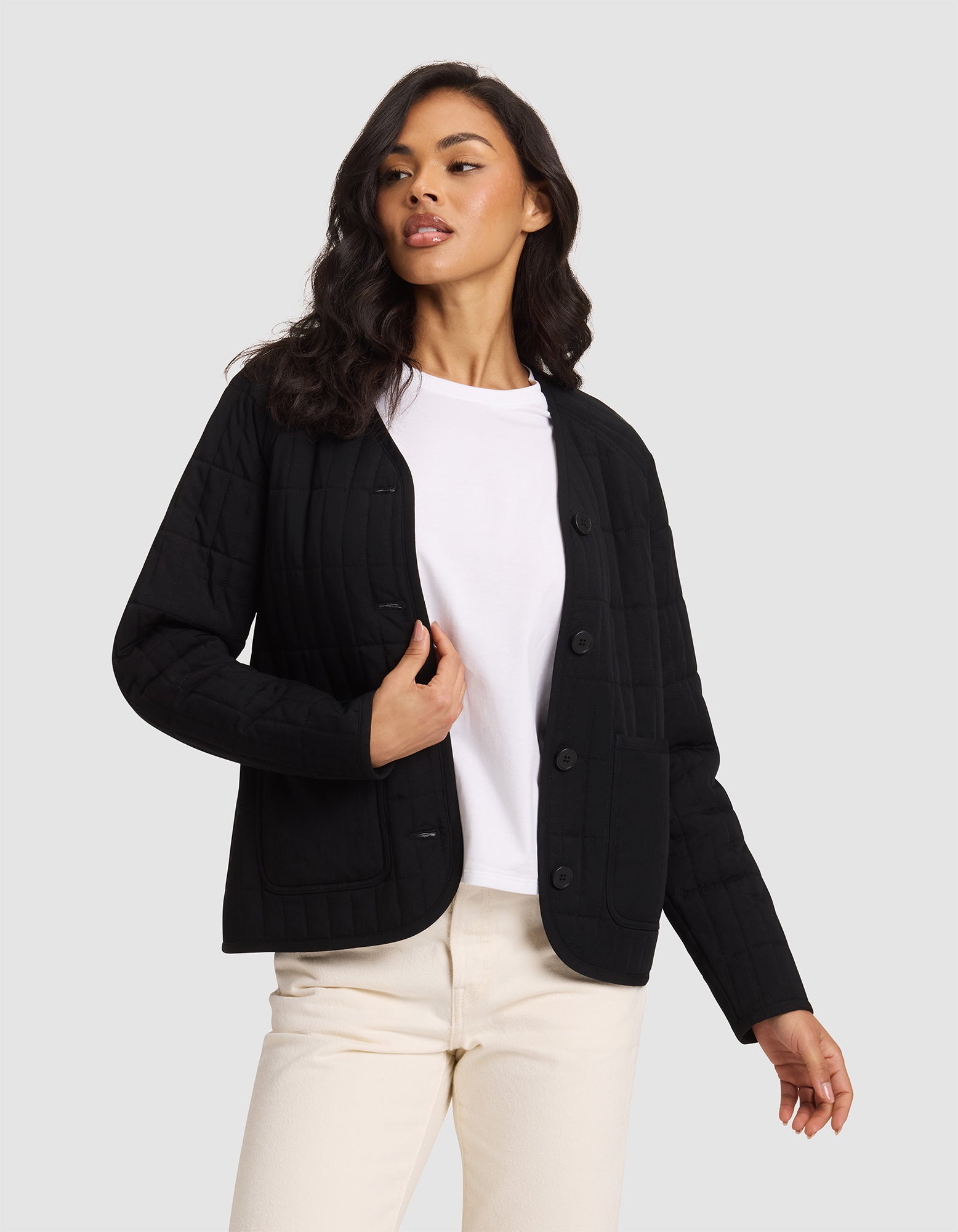 A woman with long, wavy dark hair stands against a light gray background wearing the Cozy Earth Women's Andie Quilted Jacket in black over a white t-shirt and cream-colored pants, looking slightly to the side.