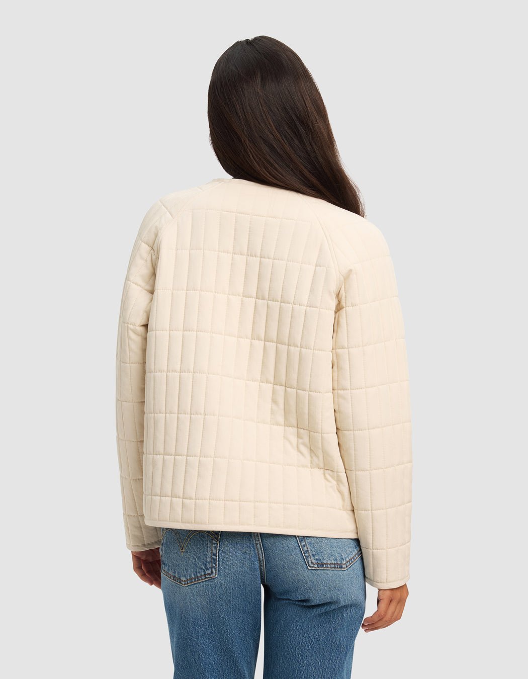 A woman with long dark hair wears the Cozy Earth Women's Andie Quilted Jacket in cream and blue jeans, standing with her back to the camera against a plain light gray background.