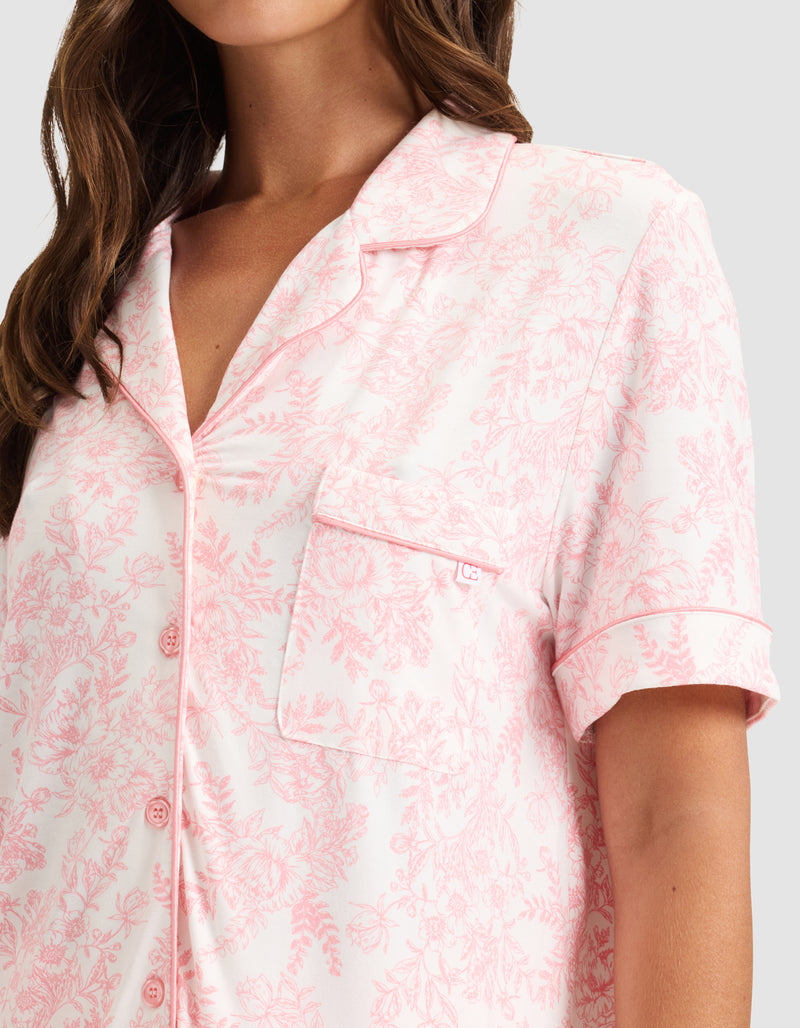 A woman wears HIDE's Women's Bamboo Stretch-Knit Short Sleeve Pajama Top in light pink with a floral pattern, notched collar, chest pocket, front buttons, and pink piping. Only her upper body is visible.