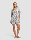 A woman with long, wavy blonde hair poses barefoot against a light gray background, wearing Cozy Earth’s Women's Bamboo Stretch-Knit Short Sleeve Pajama Set in black and white floral print shorts and matching button-up top.|Color:Jet Black Toile
