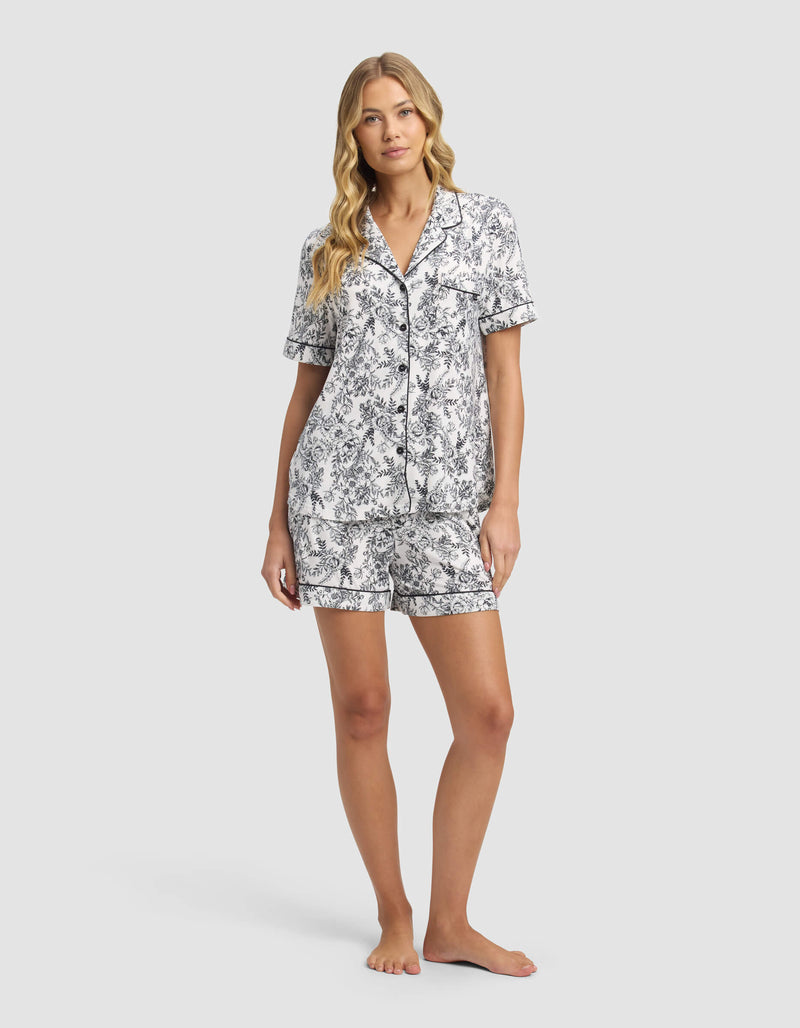 A woman with long, wavy blonde hair poses barefoot against a light gray background, wearing Cozy Earth’s Women's Bamboo Stretch-Knit Short Sleeve Pajama Set in black and white floral print shorts and matching button-up top.