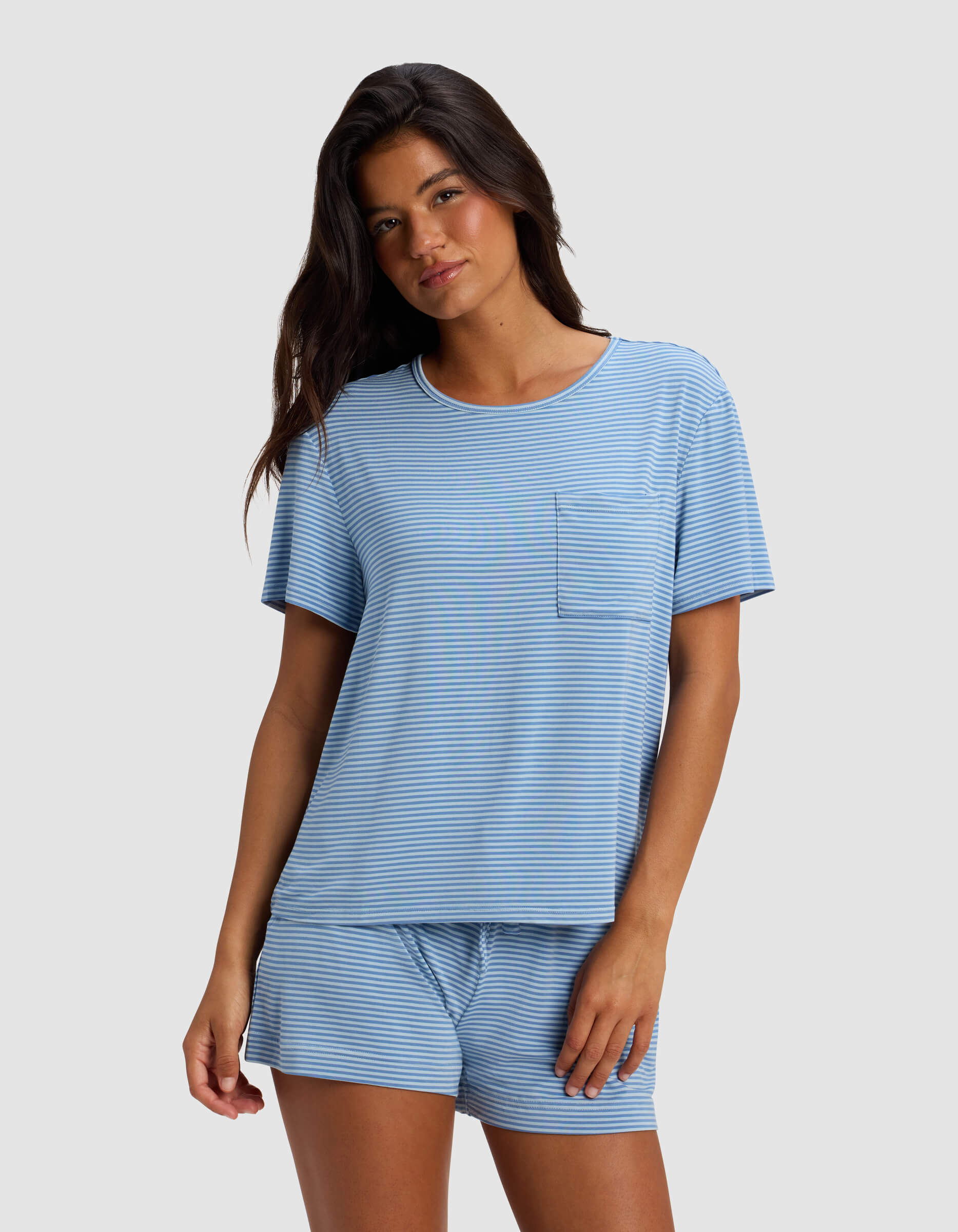 A woman wearing the Cozy Earth Women's Bamboo Stretch-Knit Lounge Tee in light blue and white stripes with a chest pocket and matching drawstring shorts stands against a plain light gray background.|Color:Sailor Mini Stripe