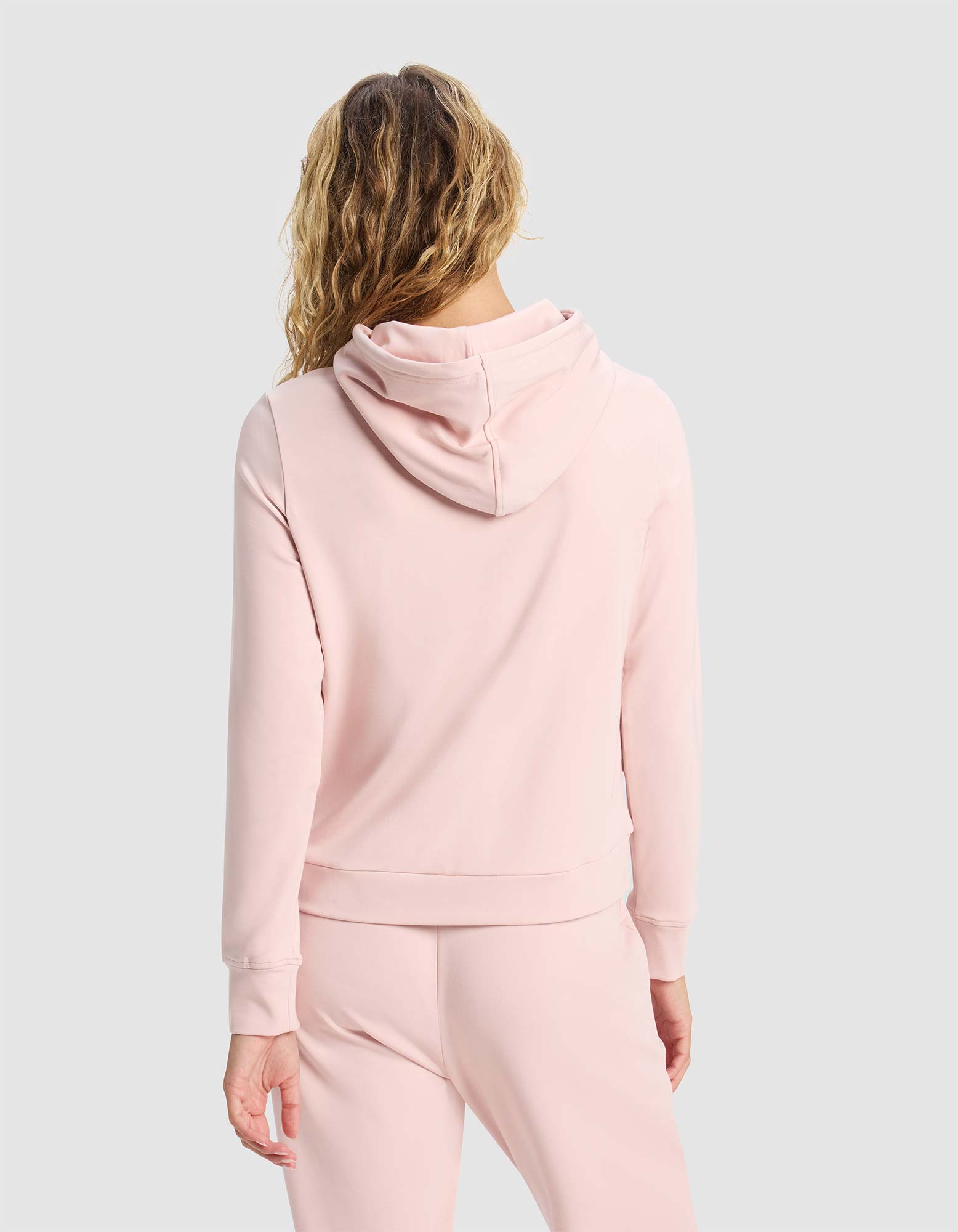A woman with wavy blonde hair is seen from the back, wearing the Cozy Earth Women's Brushed Bamboo Hoodie in light pink, paired with matching pants, against a plain light gray background. |Color:Dusty Rose