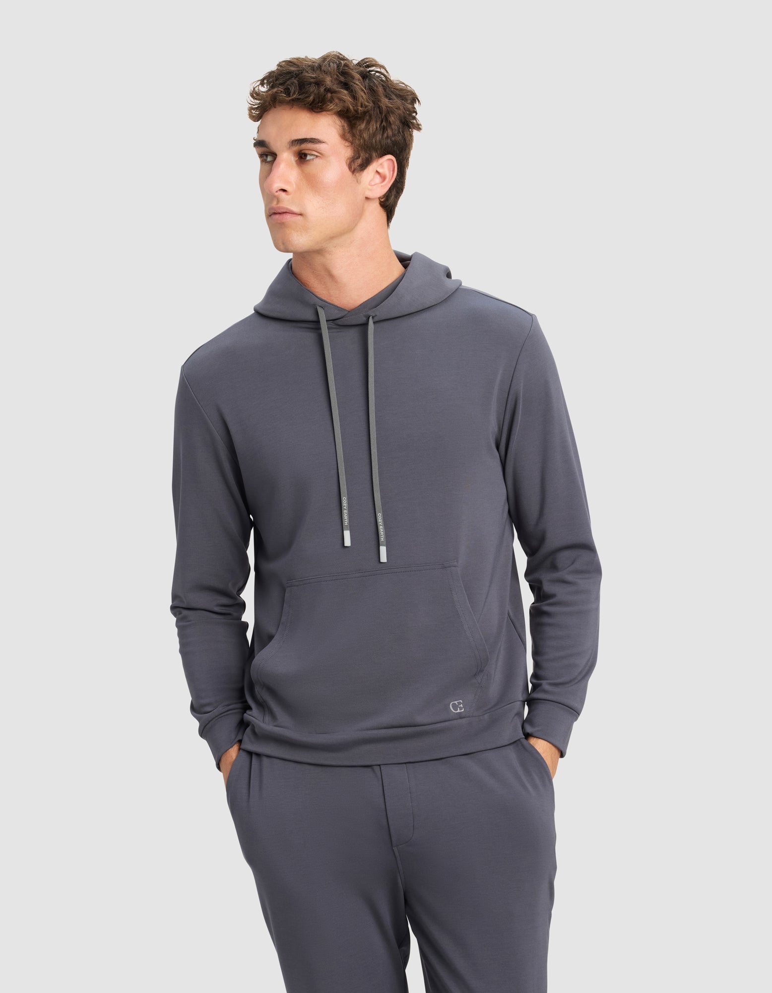 A young man models the Cozy Earth Men's Brushed Bamboo Hoodie & Jogger Set in gray, standing with his hands in his pockets and gazing slightly to the side against a plain light background.|Color:Graphite
