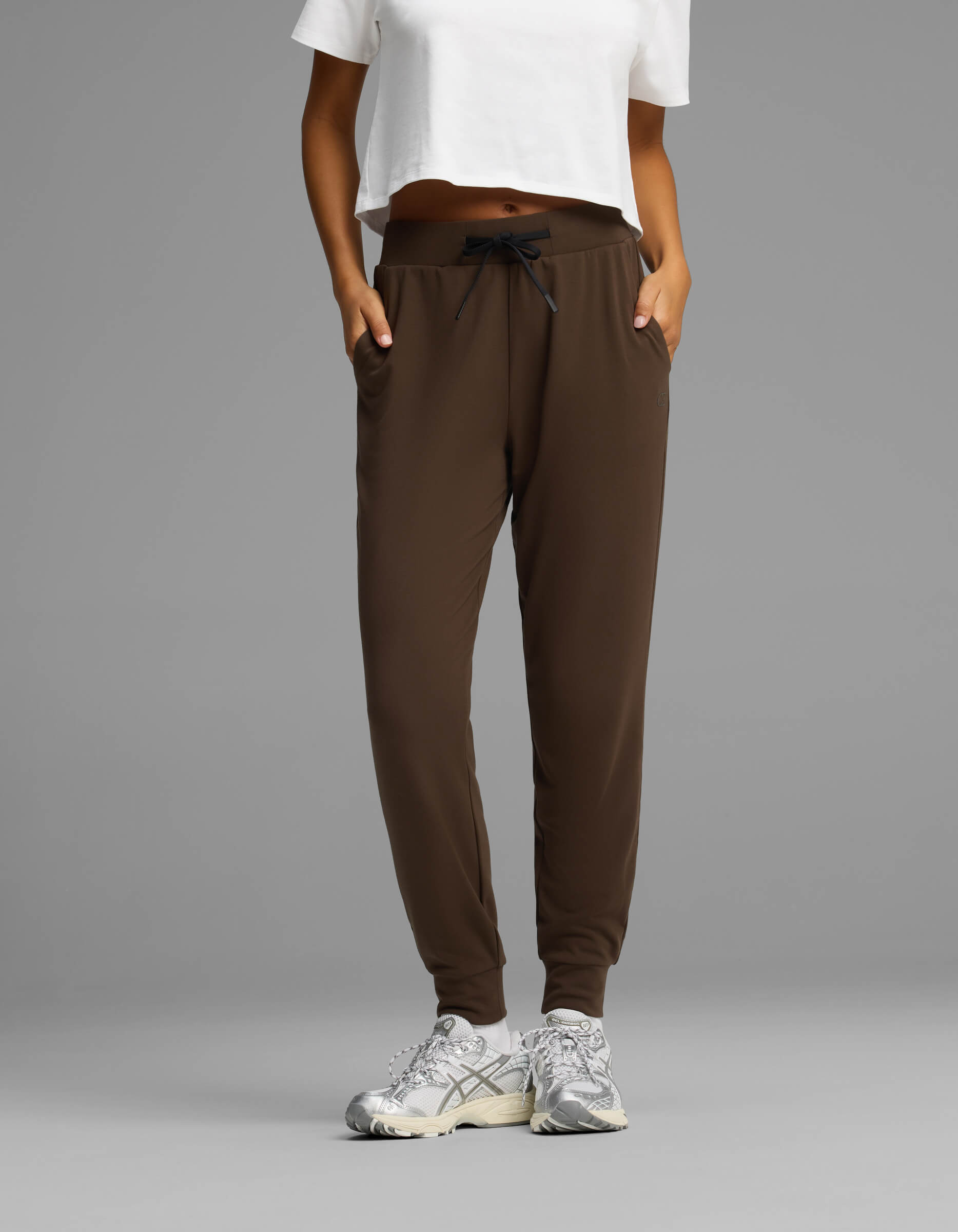 A person wearing Cozy Earth Women's Brushed Bamboo Joggers in brown, paired with a white cropped T-shirt and white sneakers, stands with hands in pockets against a plain gray background.|Color:Espresso