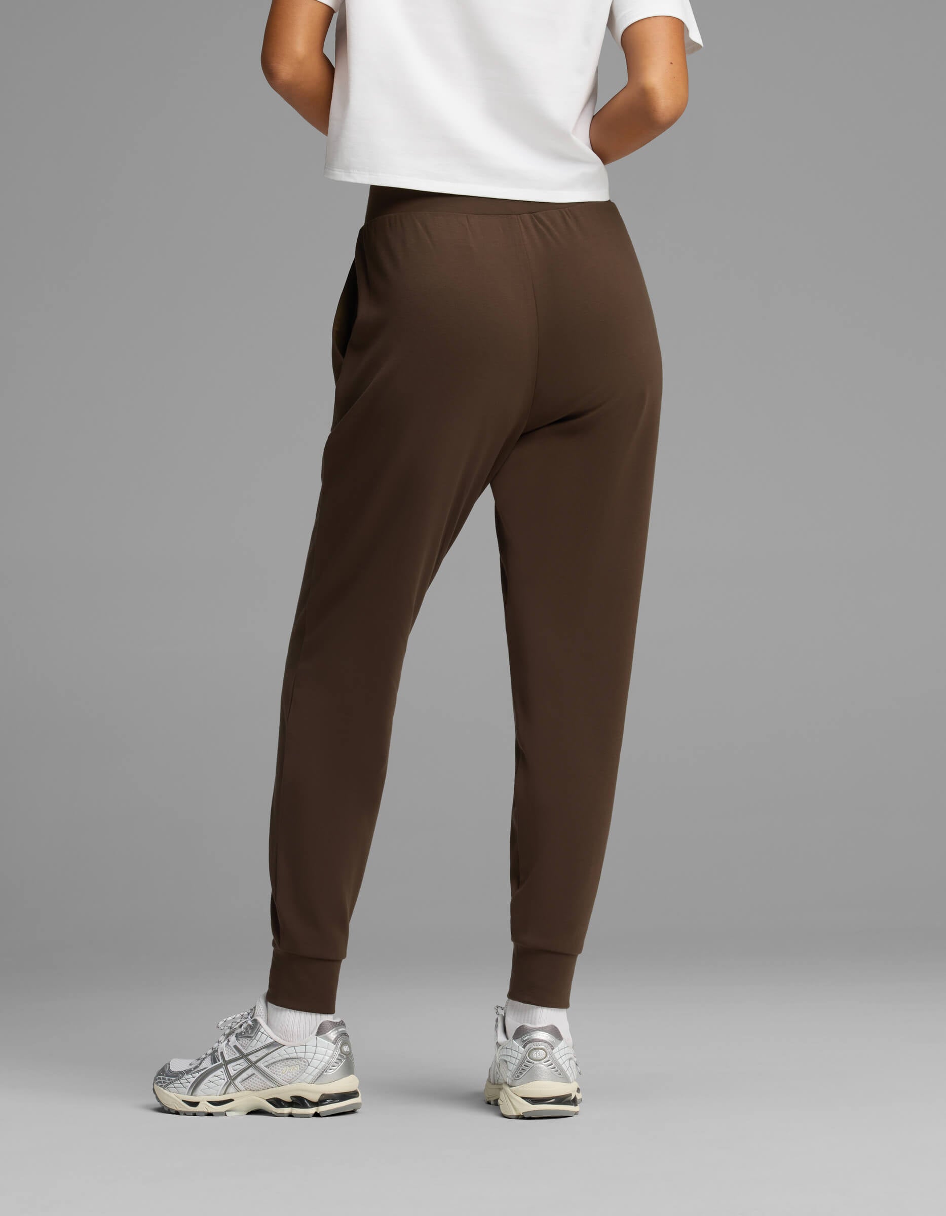 A person stands facing away against a plain gray background, wearing Cozy Earth Women's Brushed Bamboo Joggers in brown, paired with a white shirt and white sneakers.|Color:Espresso
