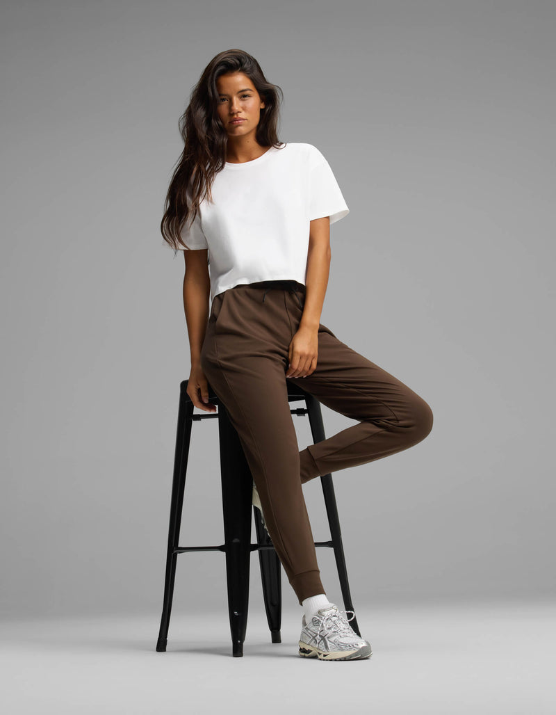 A woman models the Cozy Earth Women's Brushed Bamboo Joggers in brown, paired with a white t-shirt and white sneakers. She stands with one leg crossed, leaning on a tall black stool against a plain gray background.