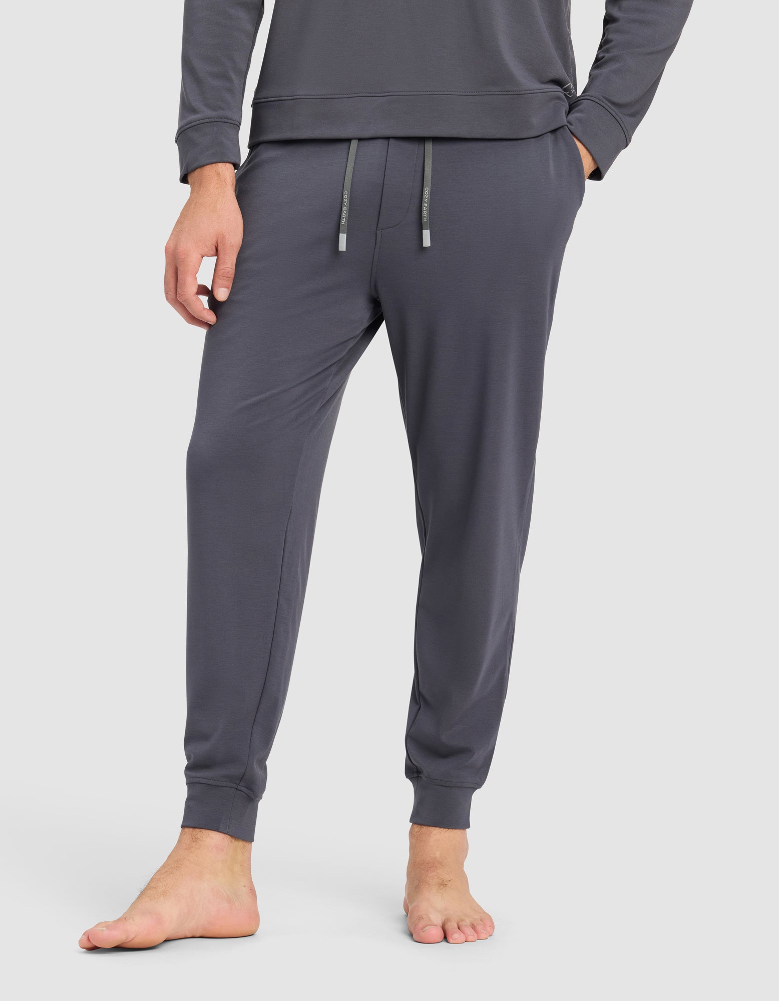 A person stands barefoot, showing only the lower half, wearing Cozy Earth's Men's Brushed Bamboo Jogger Pant in dark gray with an elastic waistband and drawstring.