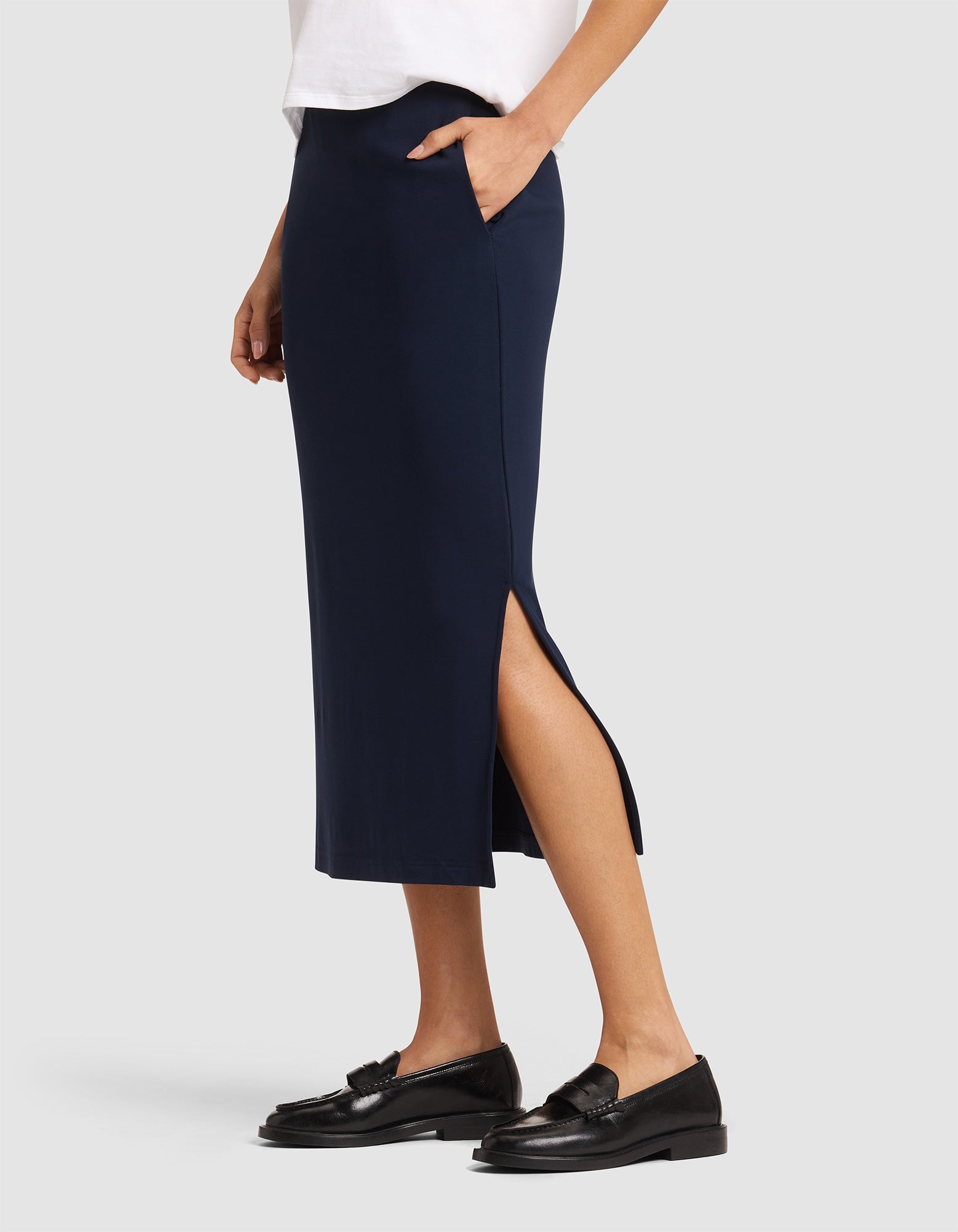 A person wearing a white top and Cozy Earth's Women's Brushed Bamboo Midi Skirt in navy blue with a side slit and pockets, paired with black loafers, stands against a plain light background.  |Color:Navy