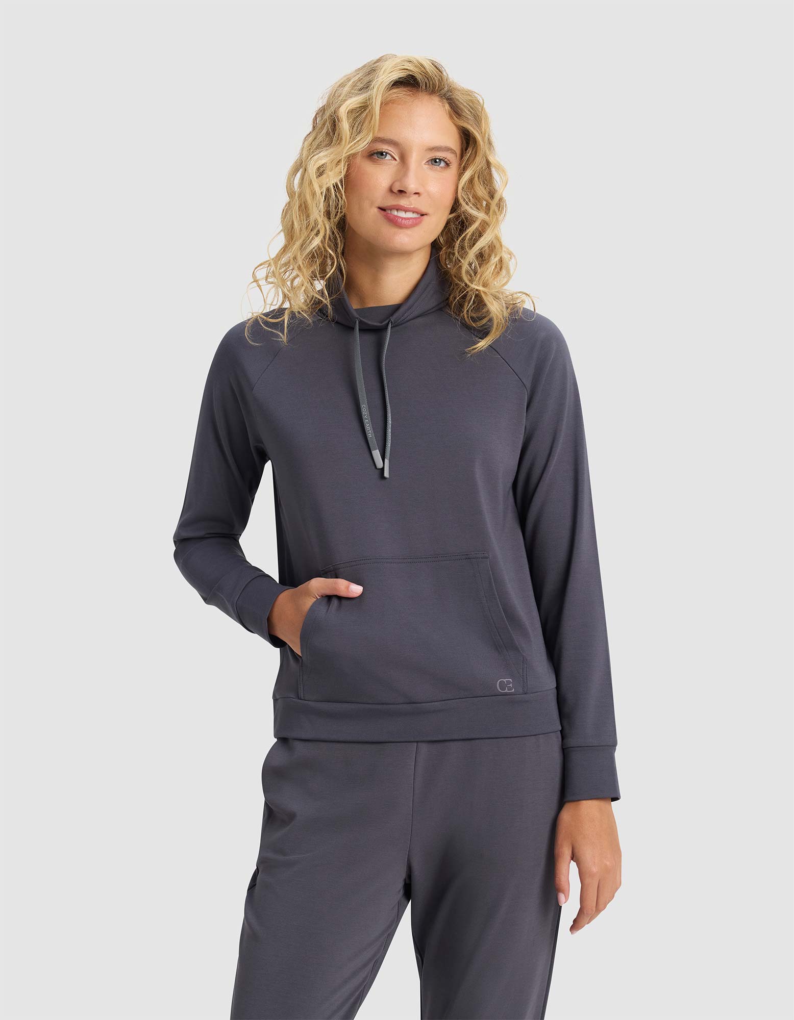 A woman with blonde curly hair wears the Cozy Earth Women's Brushed Bamboo Mock Neck in dark gray, standing against a plain light background with one hand in her pocket and a soft smile.