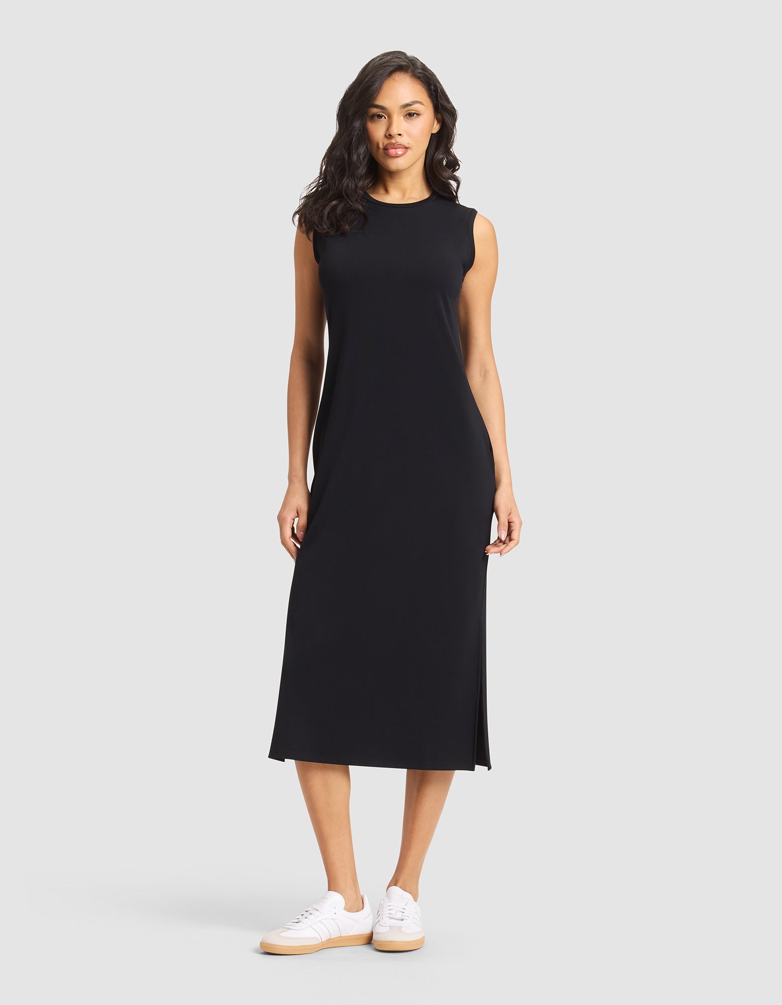 A woman with long, wavy hair and a neutral expression stands against a light gray background wearing the Cozy Earth Women's Brushed Bamboo Sleeveless Midi Dress in black, featuring side slits, paired with white sneakers. 