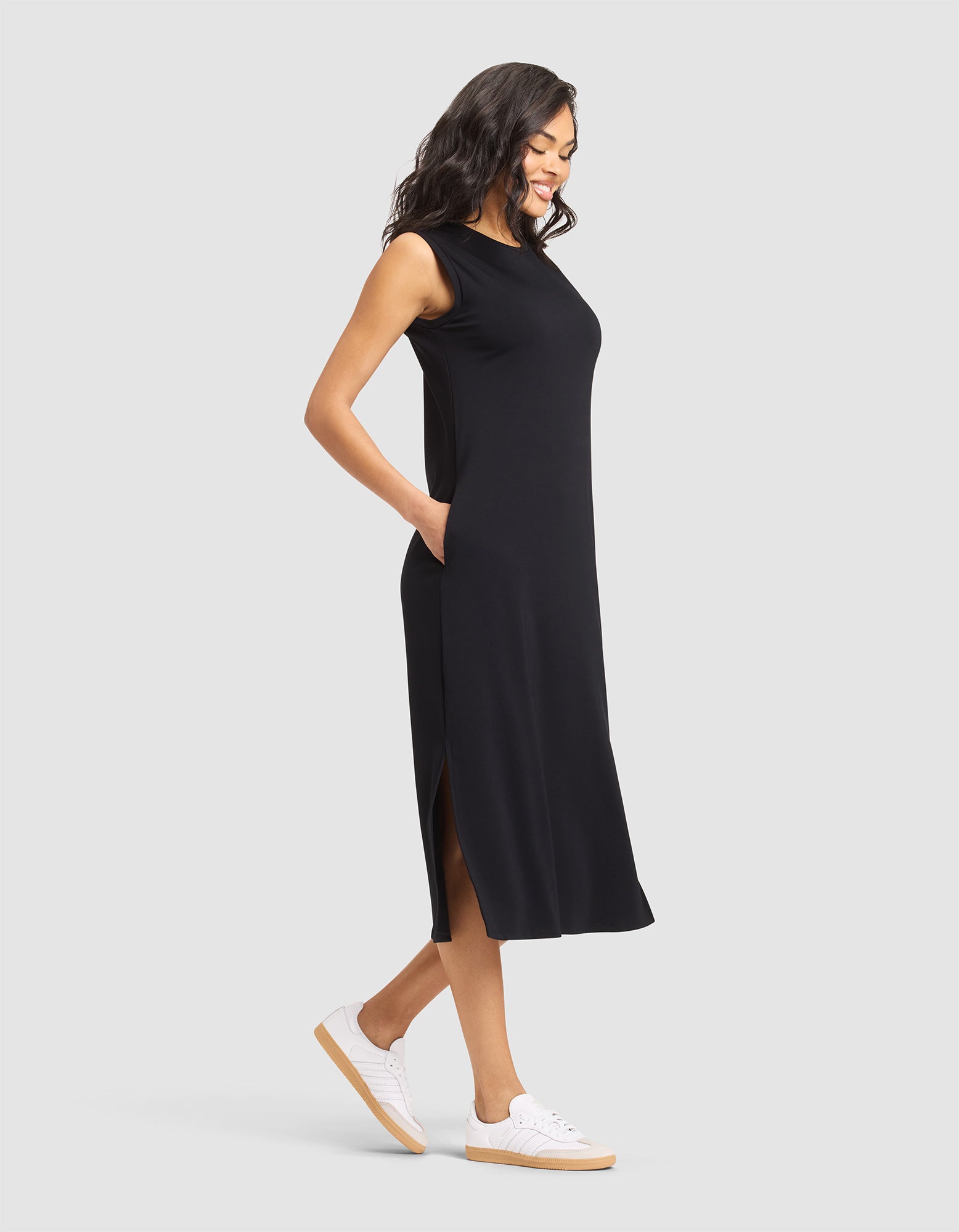 A woman smiles with her hands in her pockets, wearing the Cozy Earth Women's Brushed Bamboo Sleeveless Midi Dress and white sneakers, standing against a plain light gray background. 