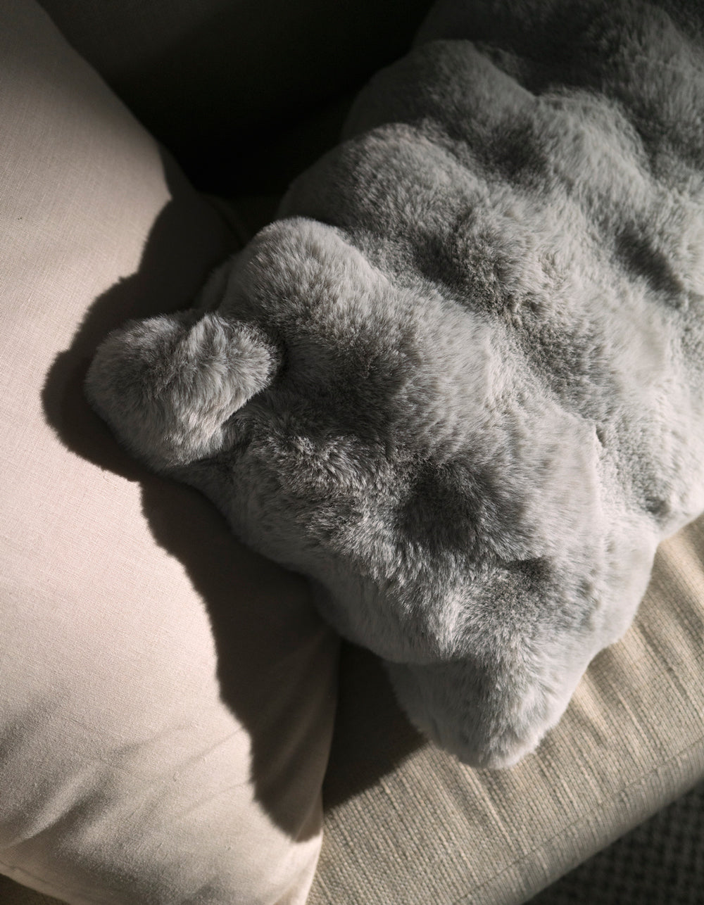 The Cozy Earth Bubble Cuddle Pillow, in soft gray faux fur, rests on a beige cushion as sunlight casts gentle shadows across its textured surface.