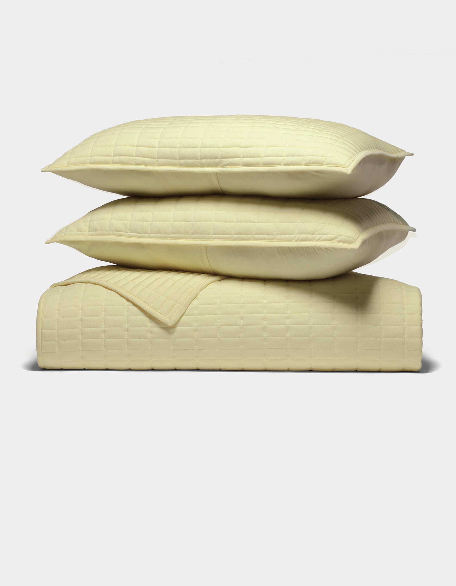 The Cozy Earth Bamboo Jersey Quilt Bundle features two pale yellow pillows stacked on a matching quilted blanket, all with a subtle square pattern, neatly arranged against a plain white background.|Color:Meringue