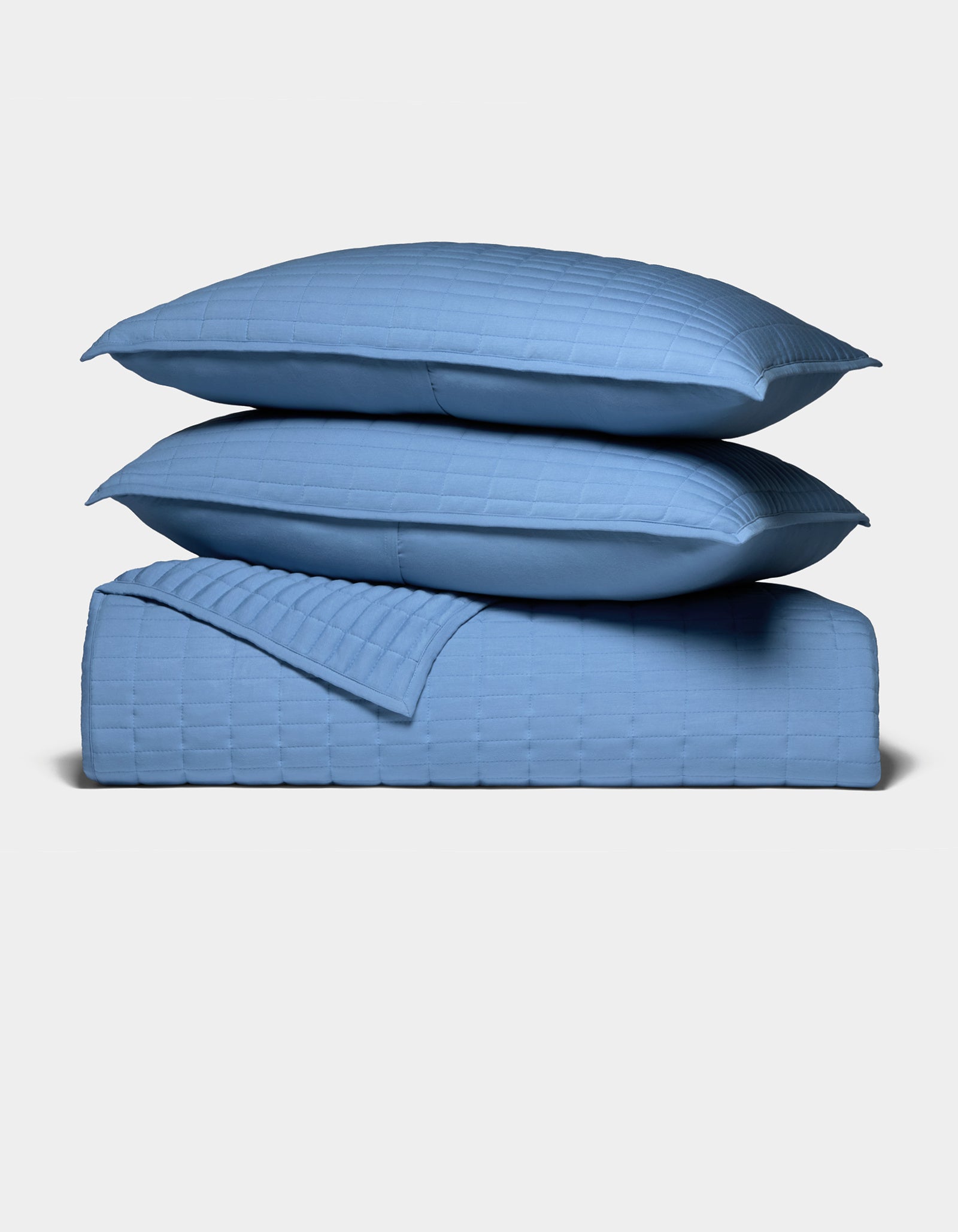 Two blue pillows rest atop a matching blue Bamboo Jersey Quilt Bundle by Cozy Earth, all showcasing a square grid pattern, set against a plain white background.|Color:Sailor