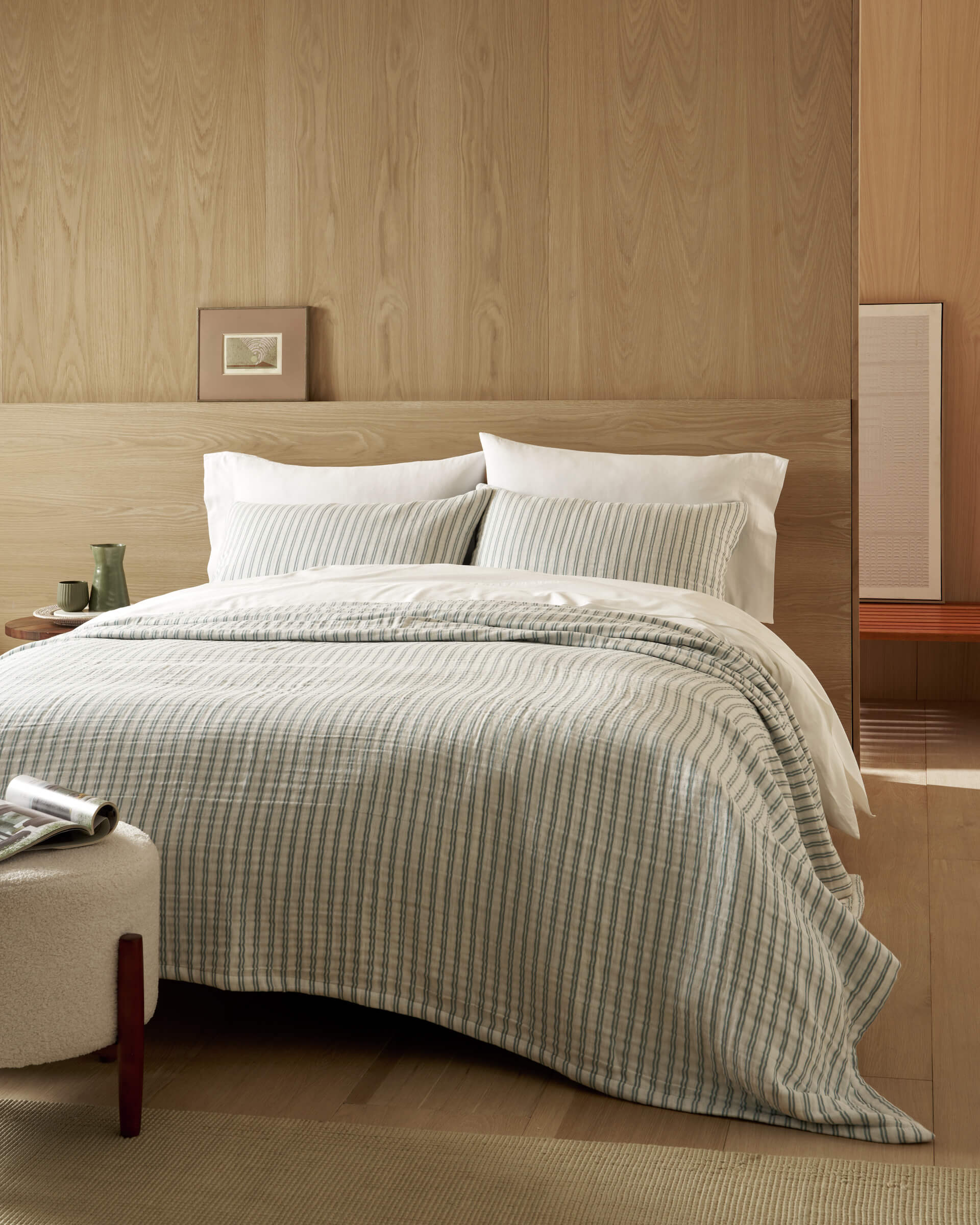 A neatly made bed features the Cozy Earth Waterfront Coverlet against a light wood-paneled wall. White pillows, a small framed picture, a green cup on the table, and a bench complete this minimalist, cozy room.